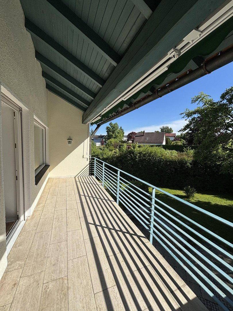 Sonniger Balkon mit Fliesenboden, blauem Geländer und Blick ins Grüne.
