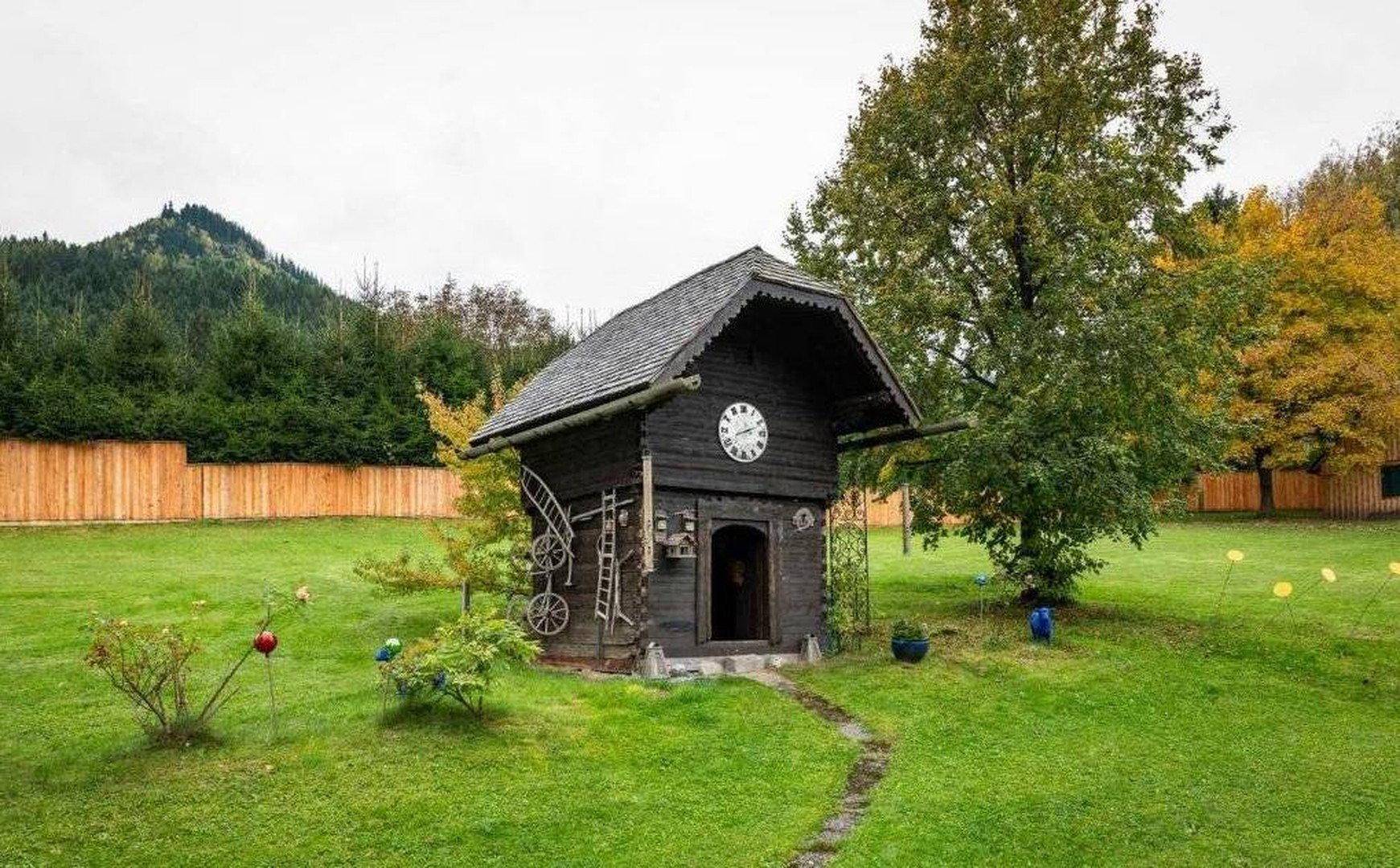 Kleines, dekoratives Gartenhaus im Stil einer Kuckucksuhr auf einer grünen Wiese.