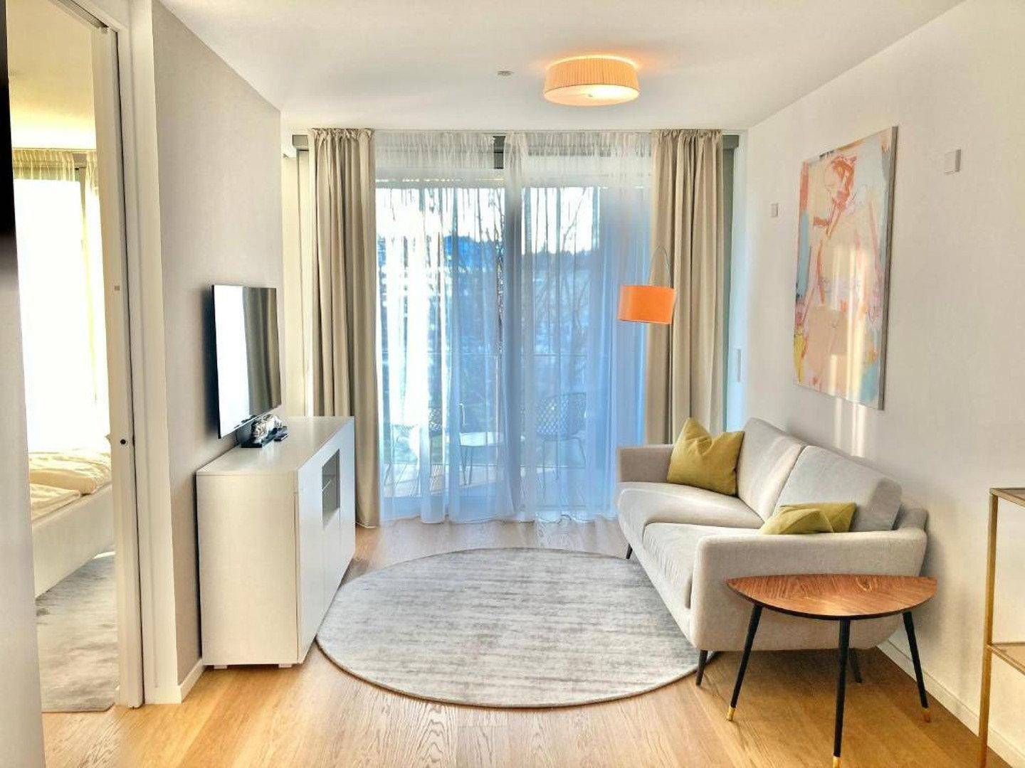 Gemütliches Wohnzimmer mit Sofa, rundem Teppich und großen Fenstern zum Balkon.