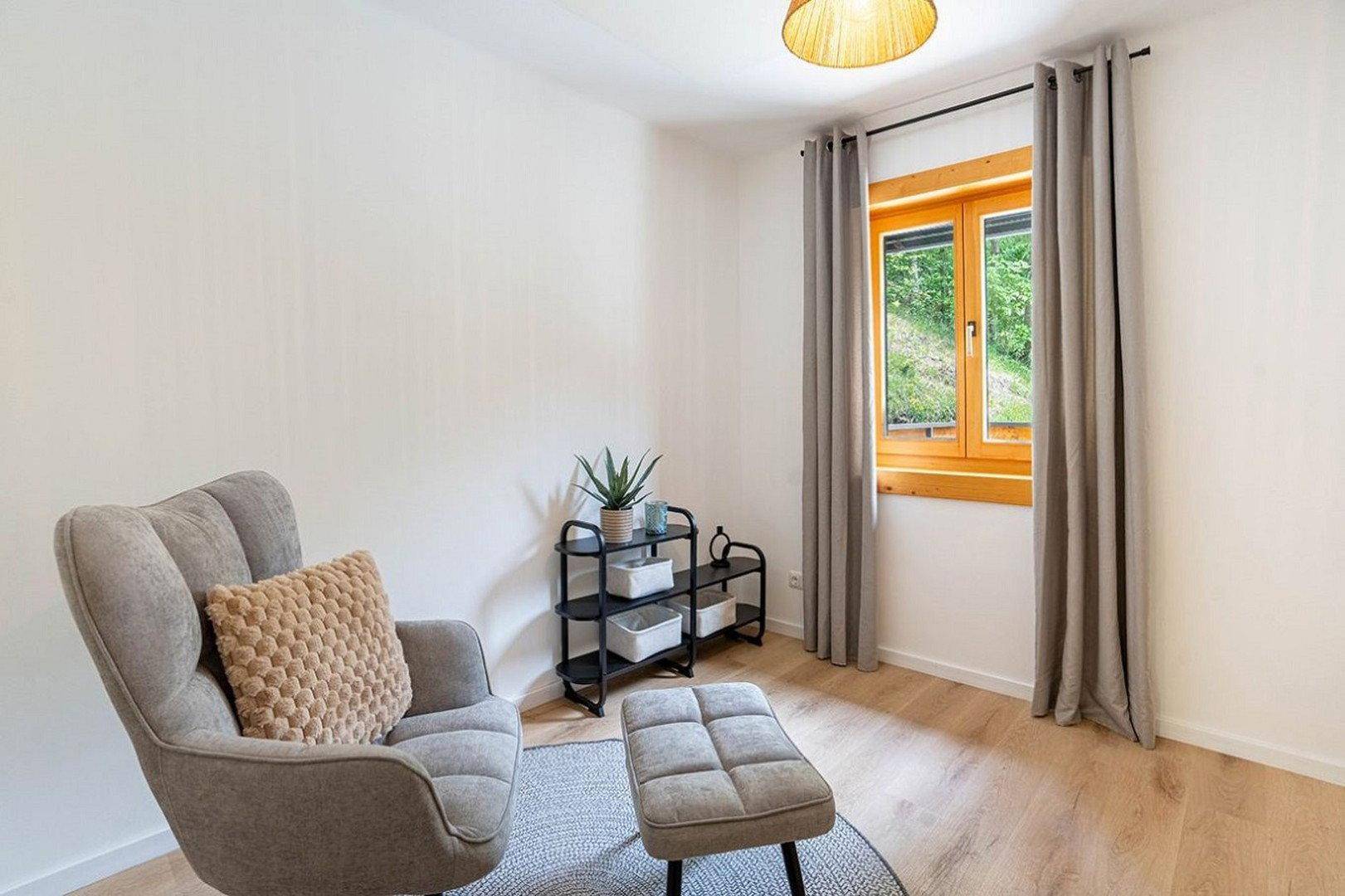 Heller Raum mit bequemem Sessel, Regal und Fenster mit Blick ins Grüne.