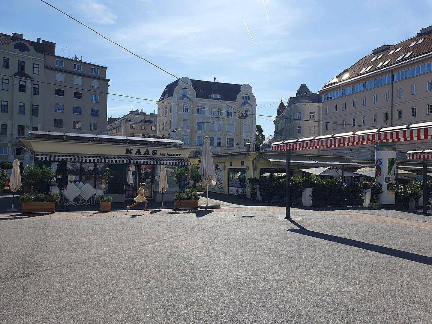 Belebte Straßenszene mit Geschäften, Cafés und Wohngebäuden im Hintergrund an einem sonnigen Tag.