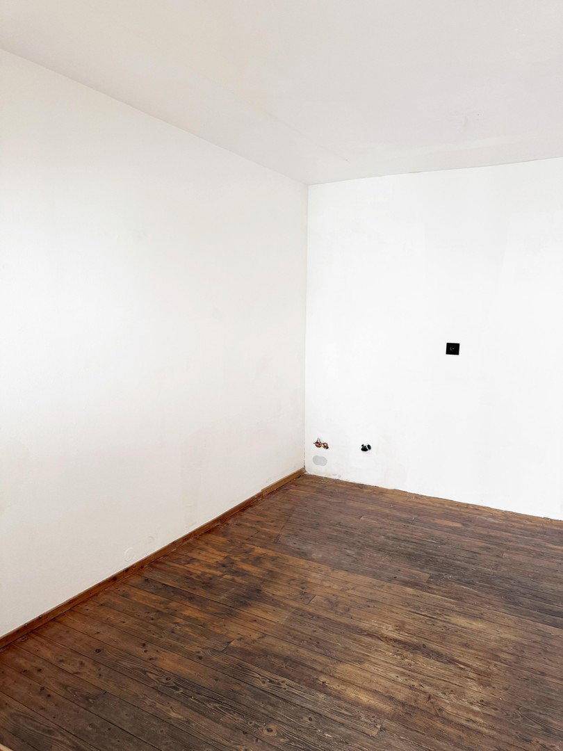 Leerer Raum mit dunklem Holzfußboden und weißen Wänden, der auf Renovierung wartet.