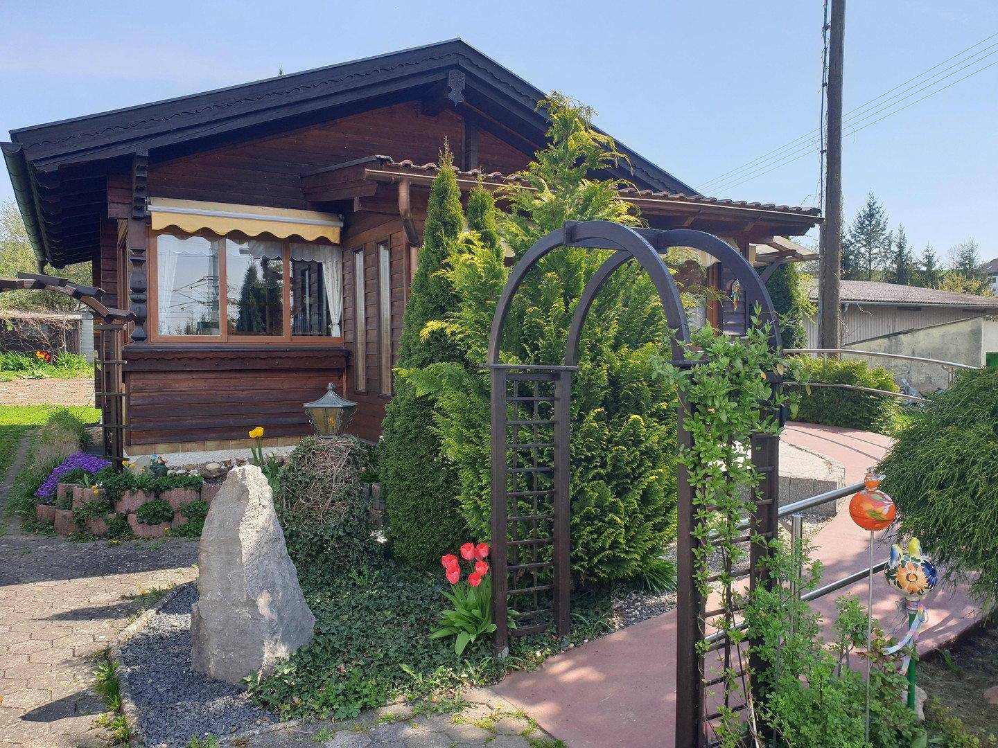 Charmantes Holzhaus mit blühenden Blumen, einem dekorativen Gartenbogen und sonnigem Wetter.