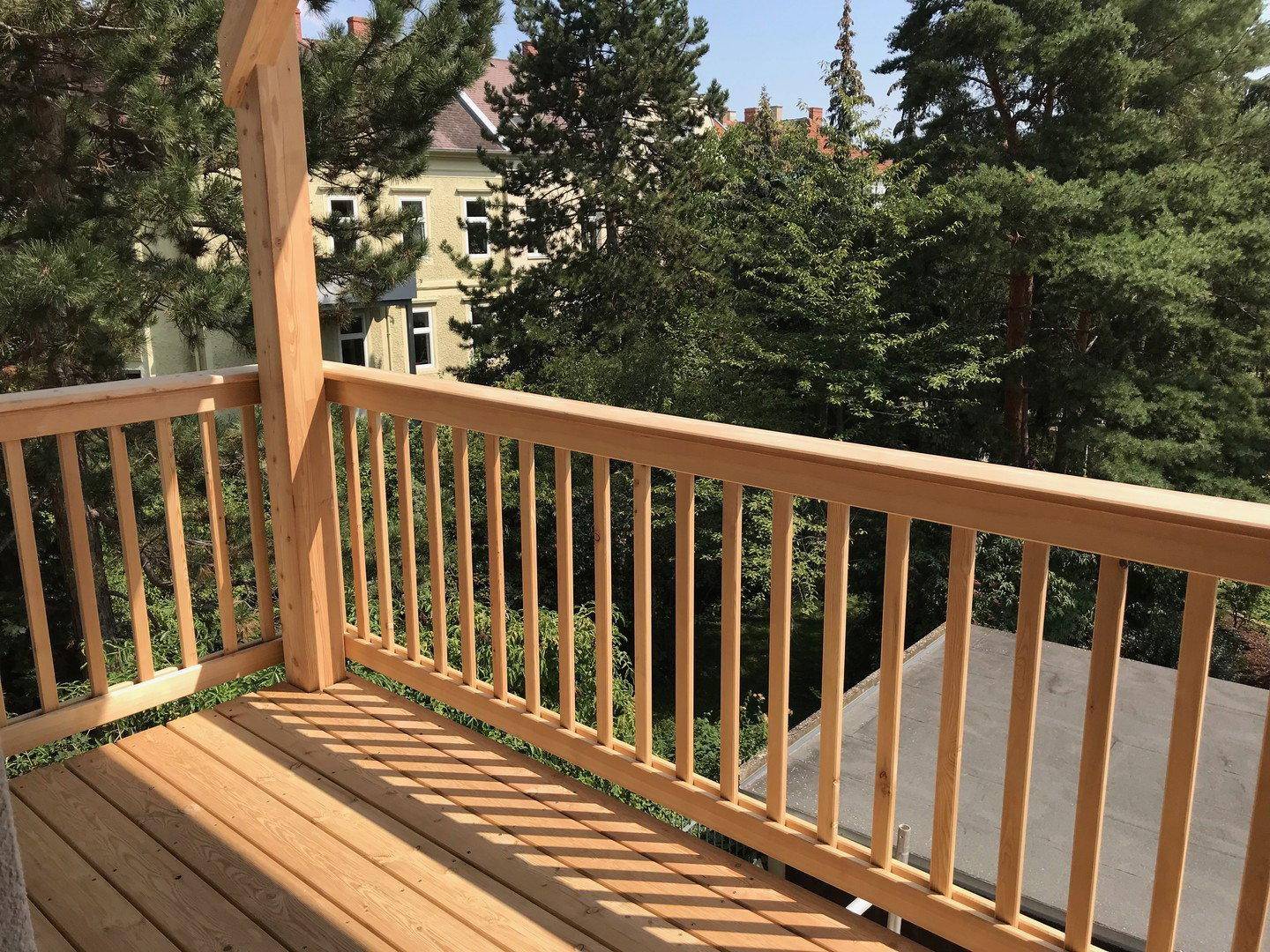 Geräumiger Balkon mit Holzgeländer und weitem Blick über grüne Bäume und umliegende Gebäude.