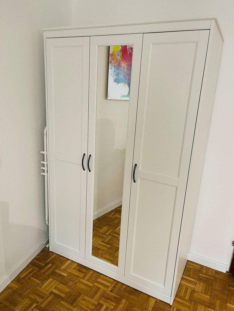 Weißer Kleiderschrank mit Spiegeltür und Holzparkettboden in einem hellen Raum.