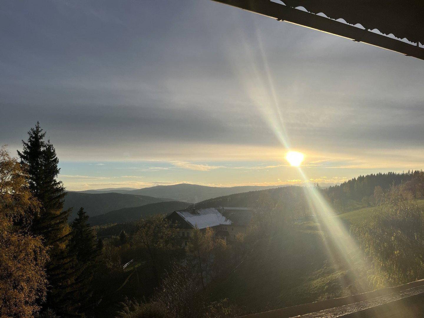 Sonnenaufgang über den Bergen und Wäldern, mit einem Haus im Tal und Schnee auf den Dächern.