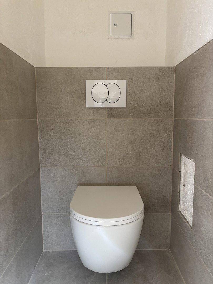 Modernes WC mit wandhängender Toilette und grauen Fliesen an Wänden und Boden.
