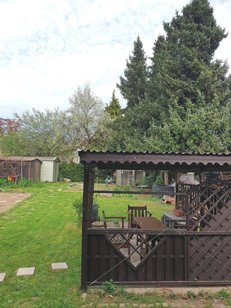 Überdachter Sitzbereich im Garten mit Holztisch und Grillplatz, umgeben von viel Grün.