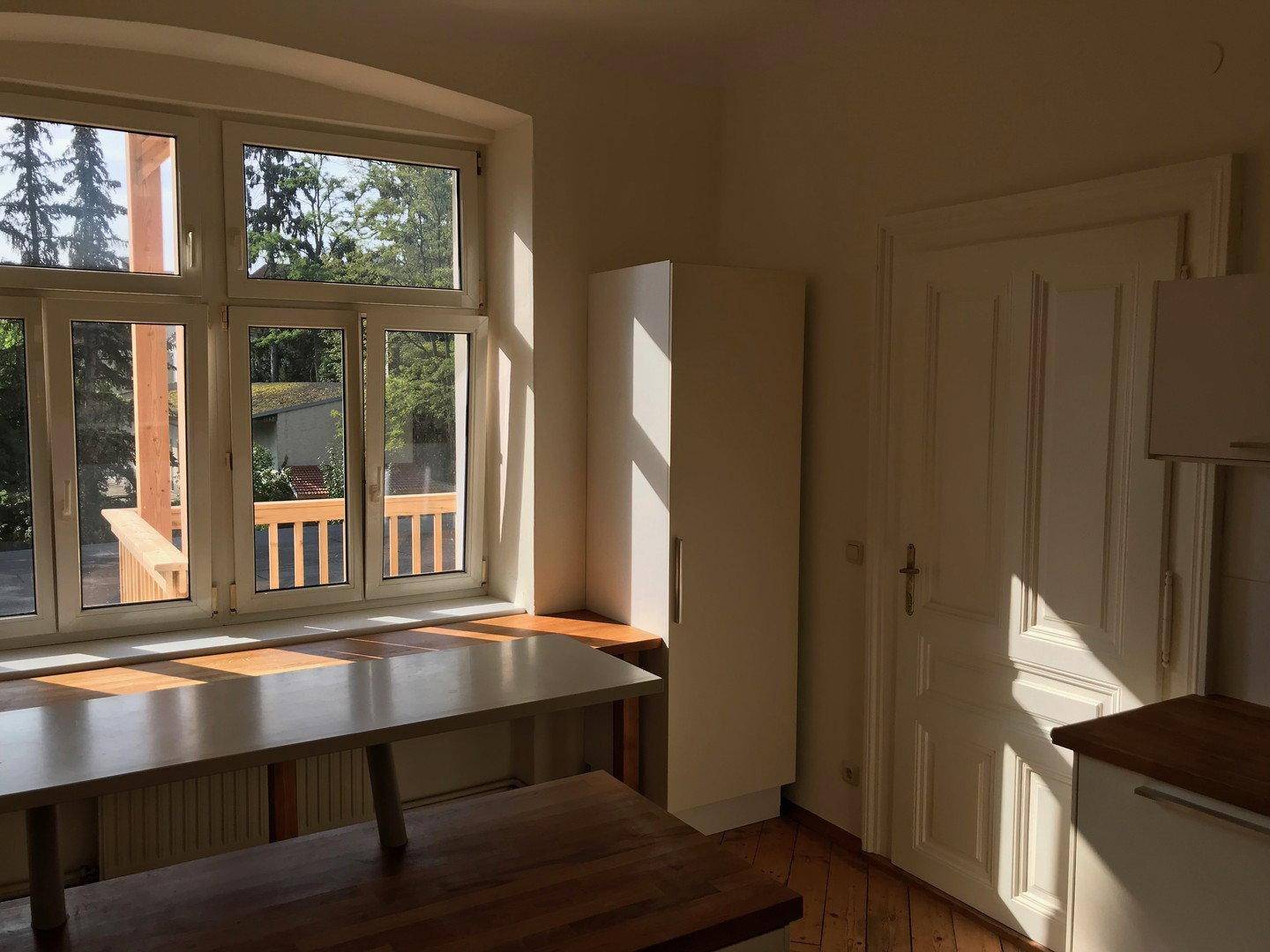 Sonniger Küchenraum mit großem Fenster, Holztisch und weißem Schrank, der viel Tageslicht hereinlässt.