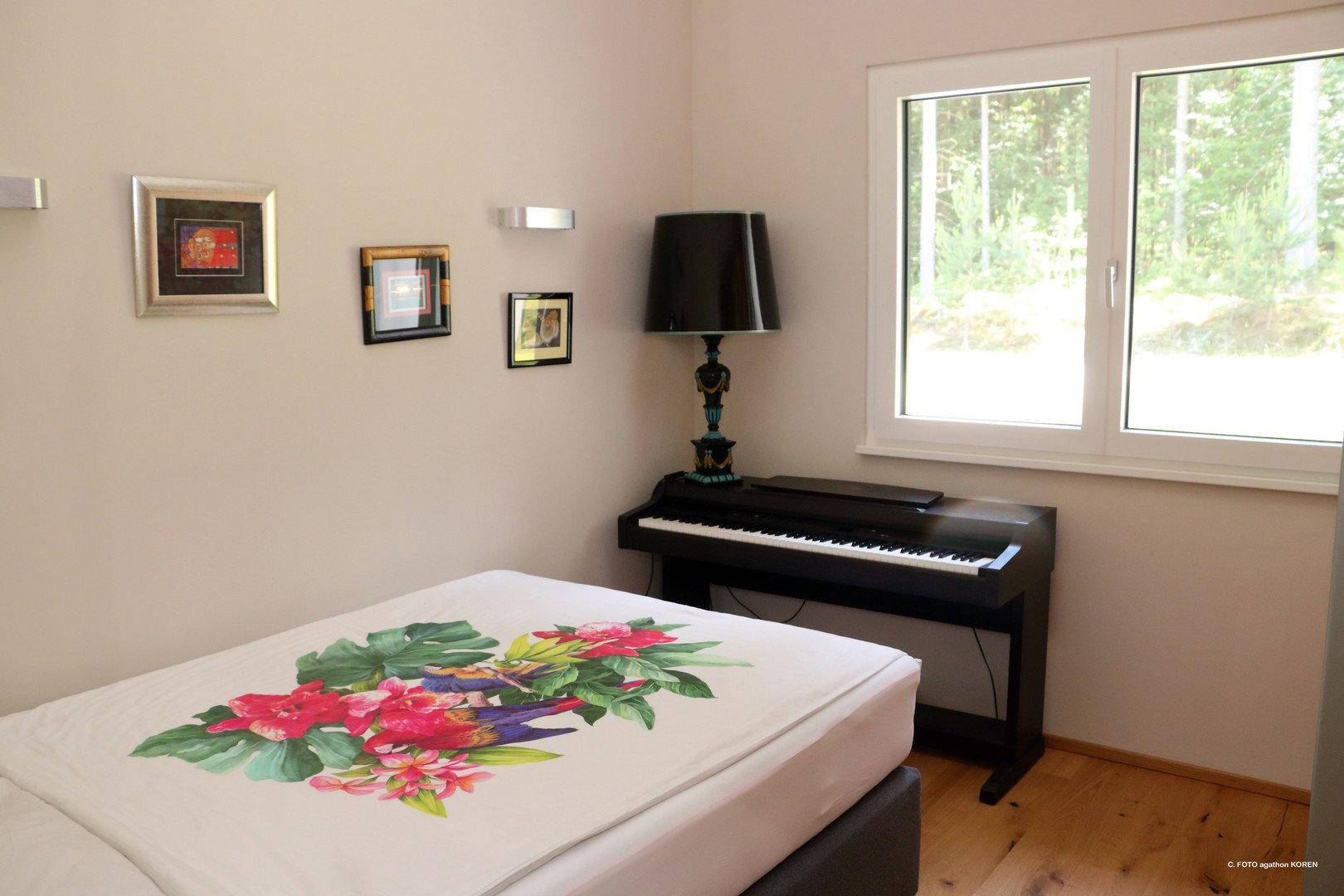 Schlafzimmer mit floral gemustertem Bett, Klavier und Fenster mit Blick ins Grüne.