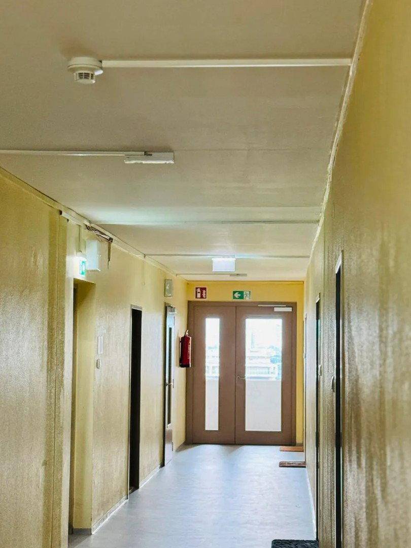 Langer Flur in einem Gebäude mit mehreren Türen und einem Rauchmelder an der Decke.