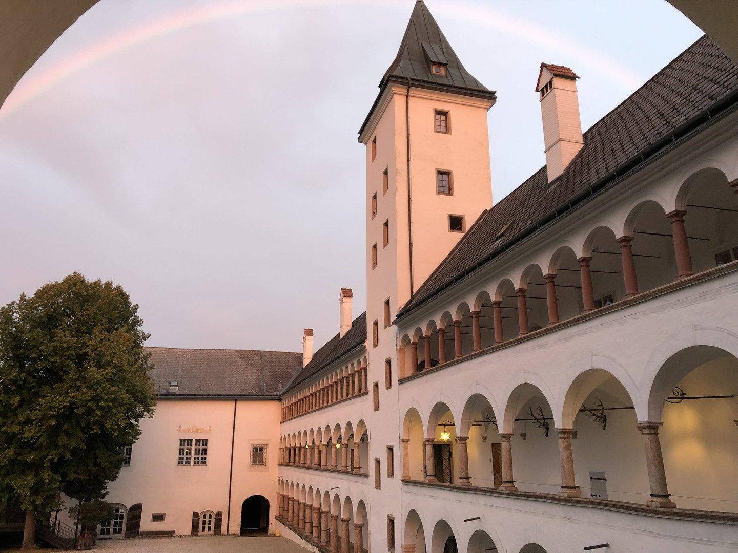 Historischer Innenhof mit Arkaden, einem Turm und einem Regenbogen am Himmel.