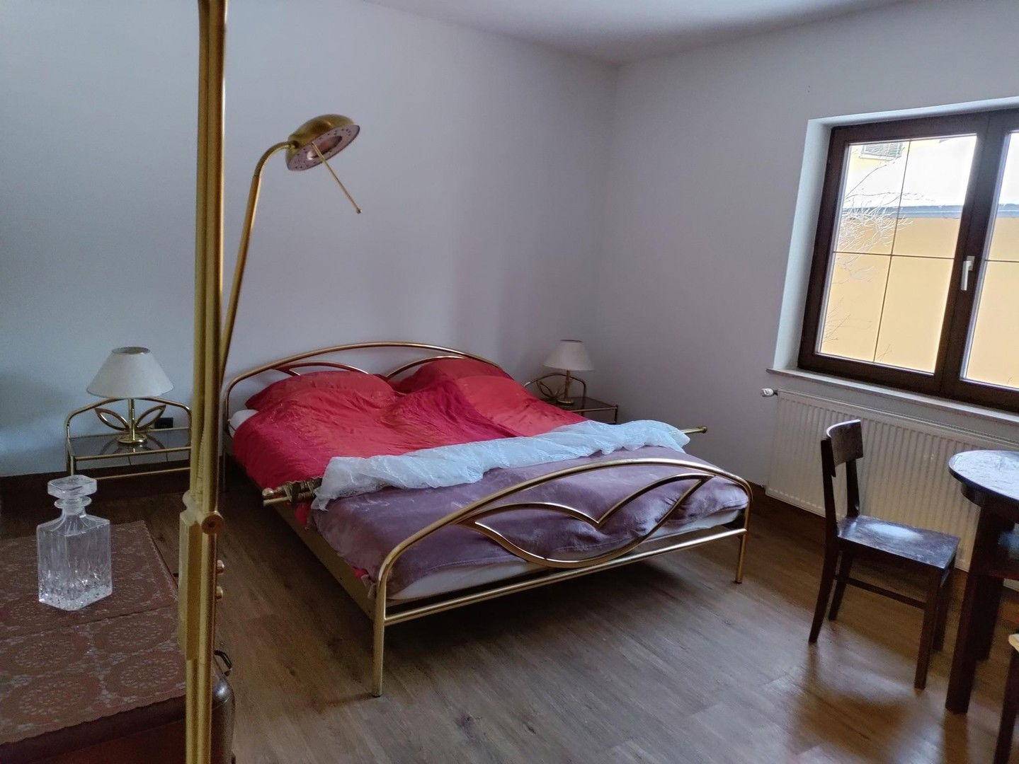 Schlafzimmer mit goldenem Metallbett, Holzboden und Fenster mit Blick nach draußen.