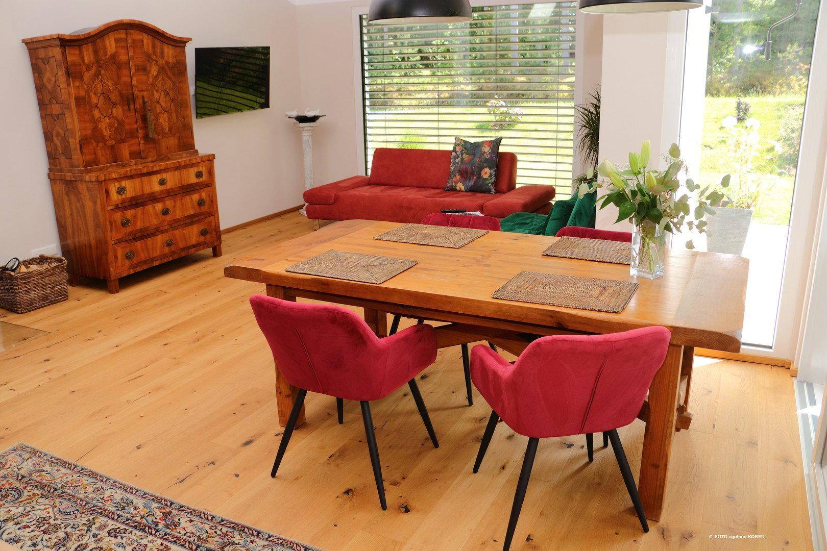 Heller Wohn- und Essbereich mit Holzboden, großem Esstisch und rotem Sofa, mit Blick ins Grüne.
