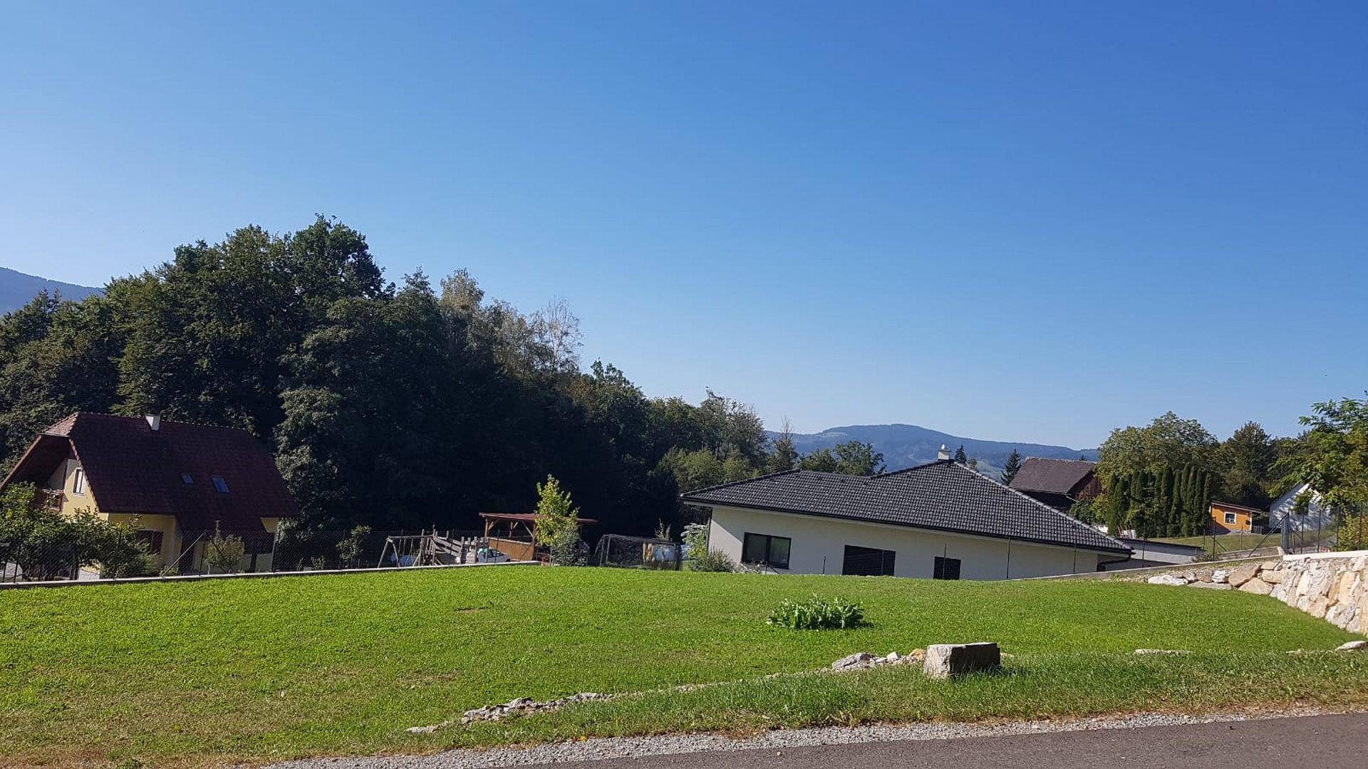 Neubauhaus mit gepflegtem Rasen und Blick auf bewaldete Hänge unter strahlend blauem Himmel.