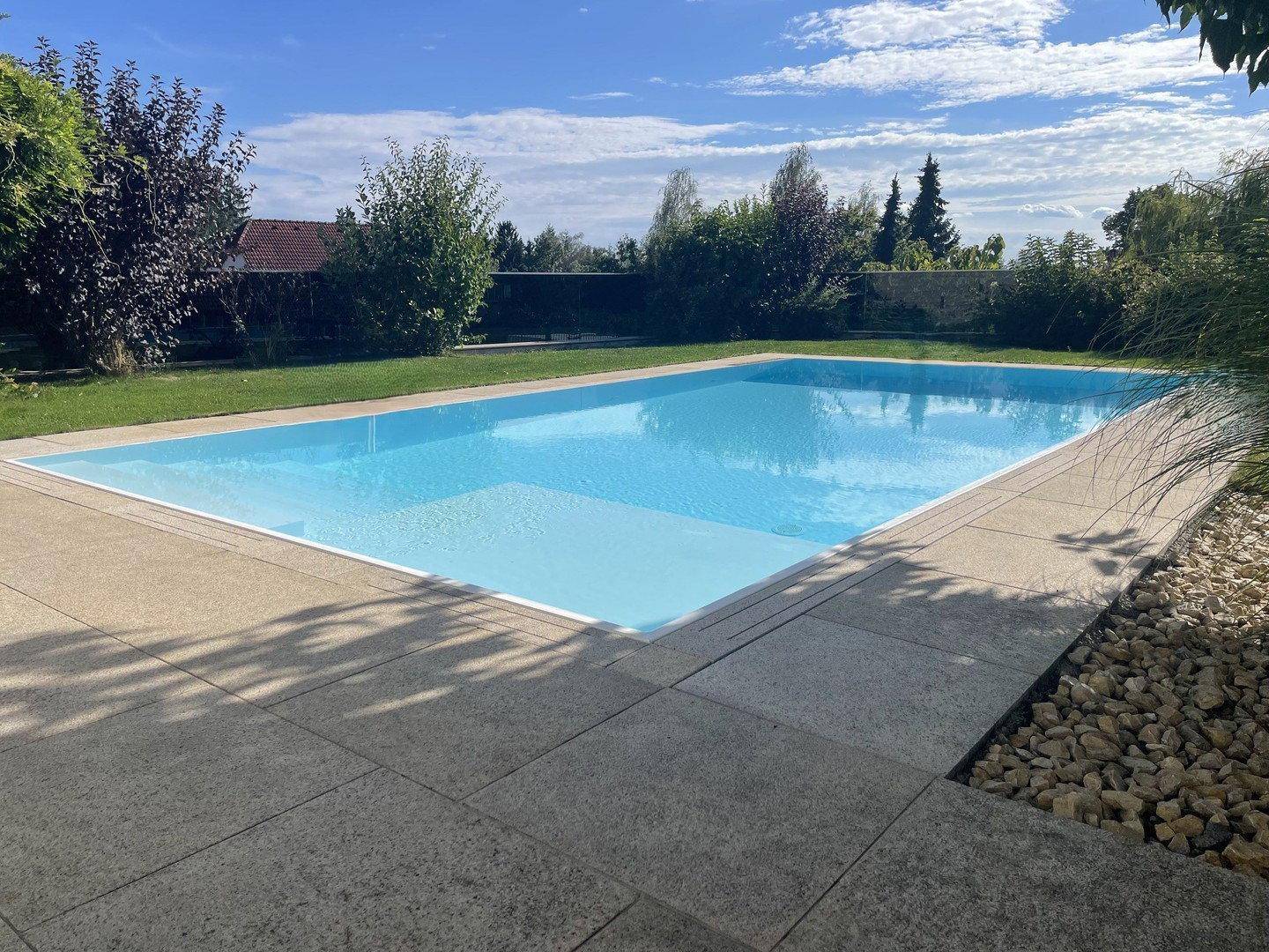 Großer Außenpool mit gepflasterter Terrasse, umgeben von Rasen und Bäumen an einem sonnigen Tag.