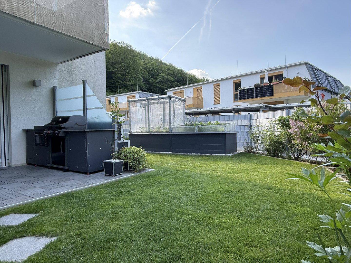 Sonniger Garten mit Terrasse, Grill und Hochbeeten für den Anbau von frischem Gemüse.