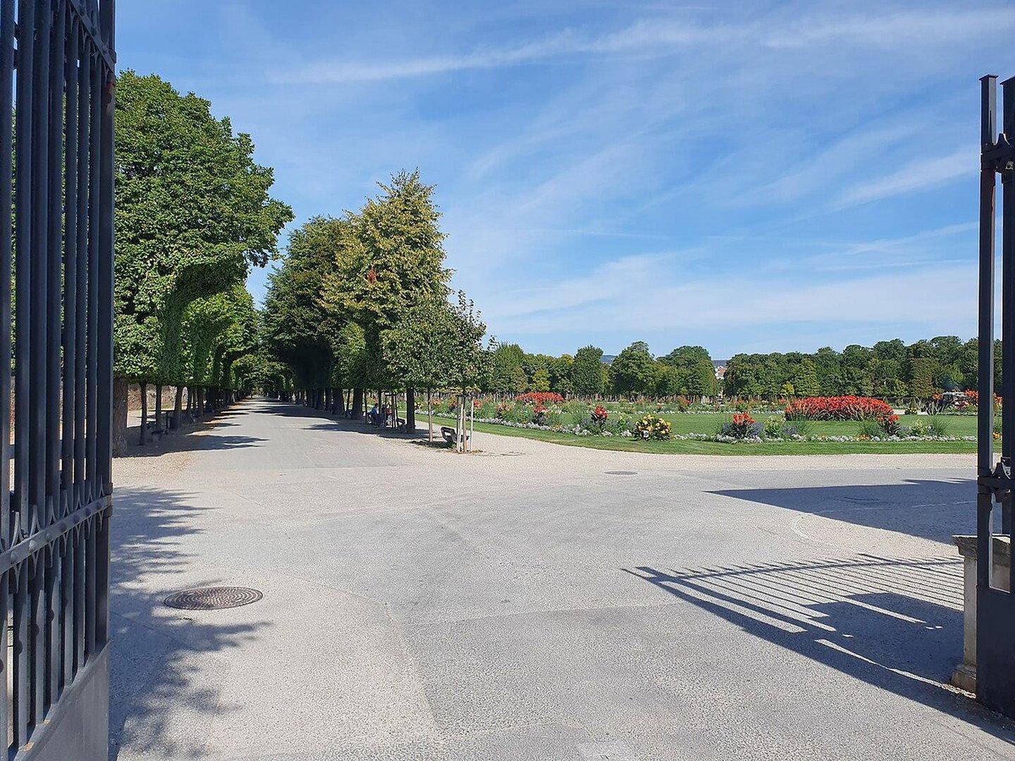 Weitläufiger Park mit langen Wegen, alten Bäumen und Blumenbeeten unter blauem Himmel.