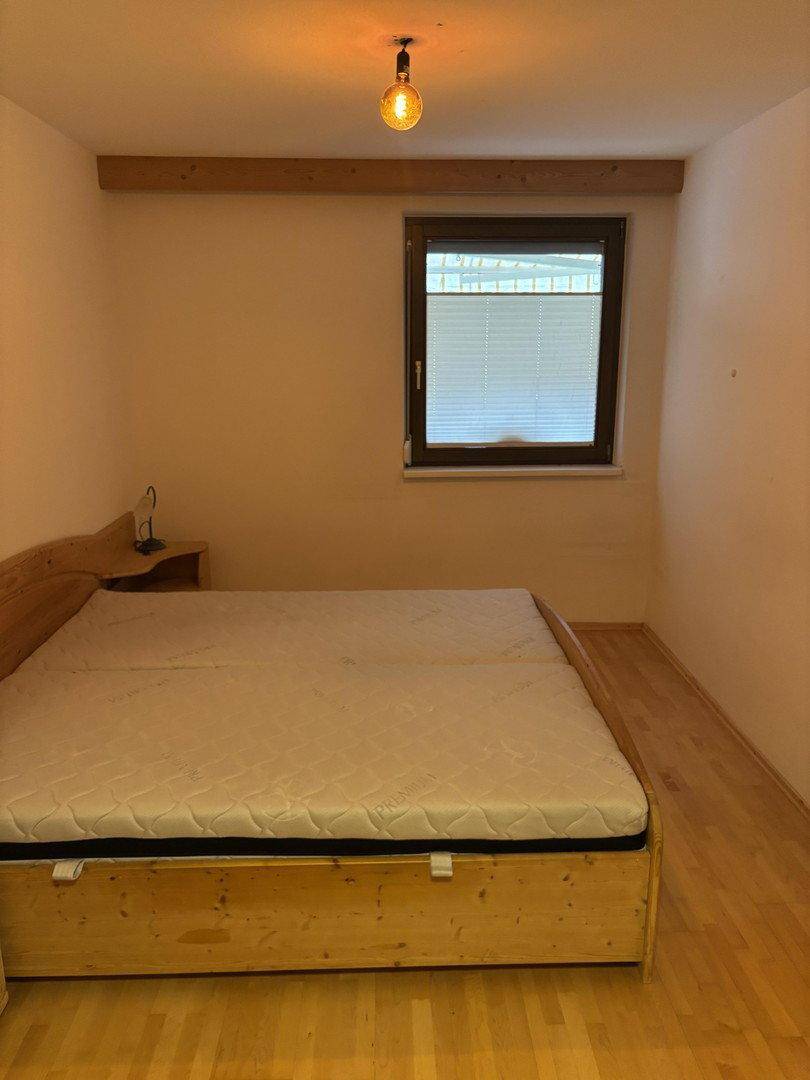 Schlafzimmer mit einem Holzbettrahmen, einem Fenster und hellen Wänden, einfach eingerichtet.