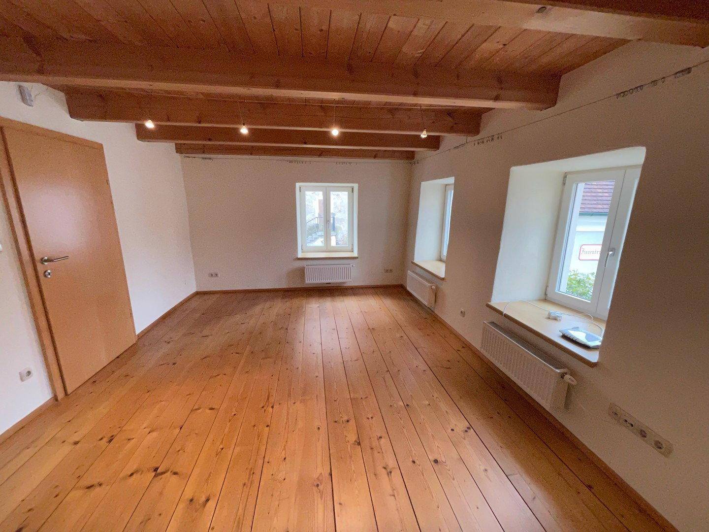 Lichtdurchflutetes Wohnzimmer mit Holzdielenboden, sichtbaren Holzbalken und Heizkörpern unter den Fenstern.