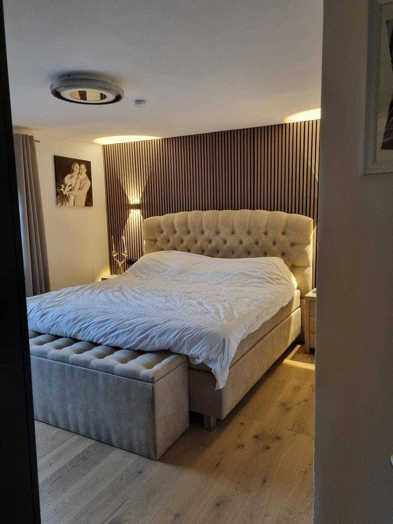 Gemütliches Schlafzimmer mit großem Polsterbett, Holzwandpaneelen und indirekter Beleuchtung.