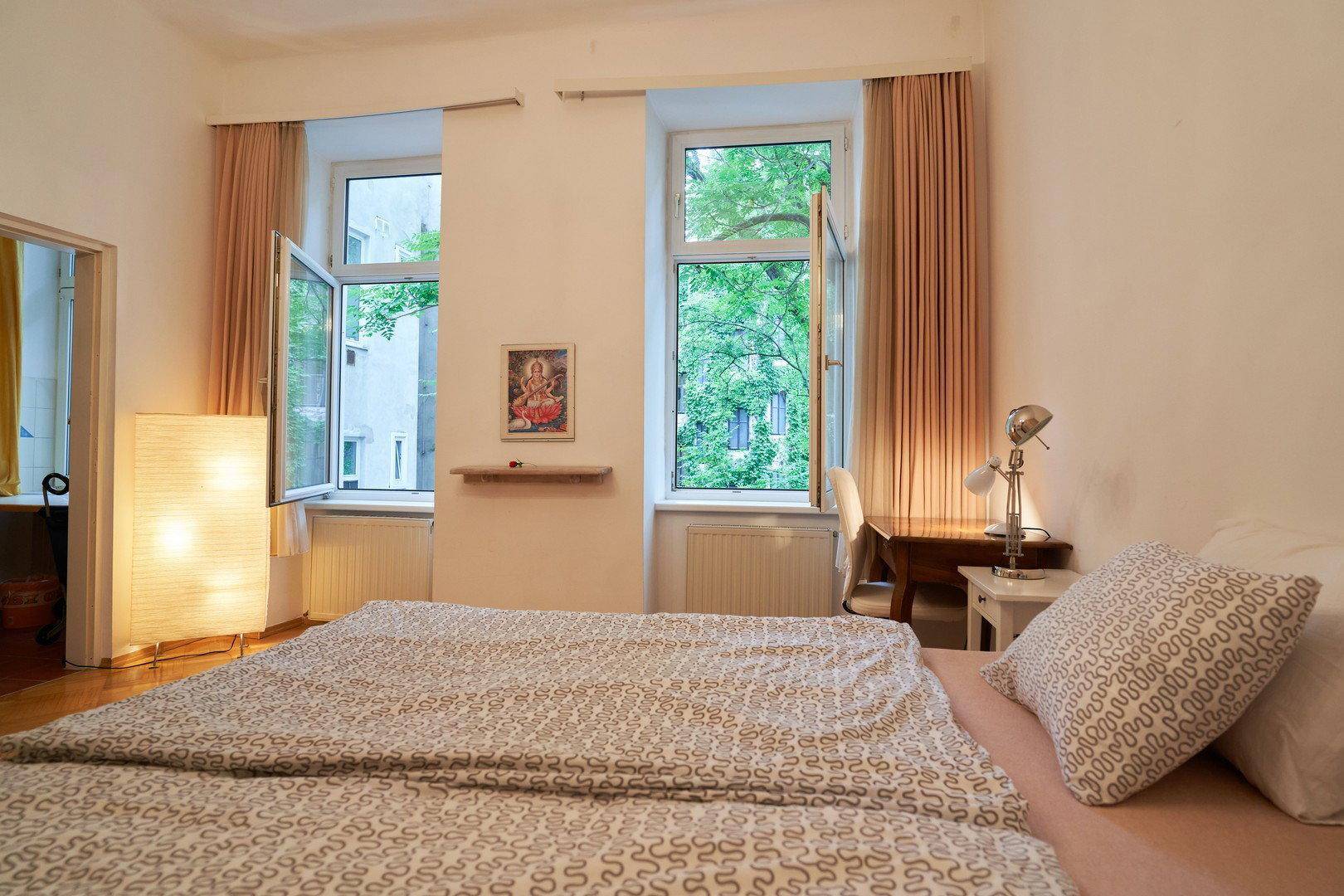 Schlafzimmer mit geöffneten Fenstern, die einen Blick ins Grüne bieten, und einem Arbeitsbereich.