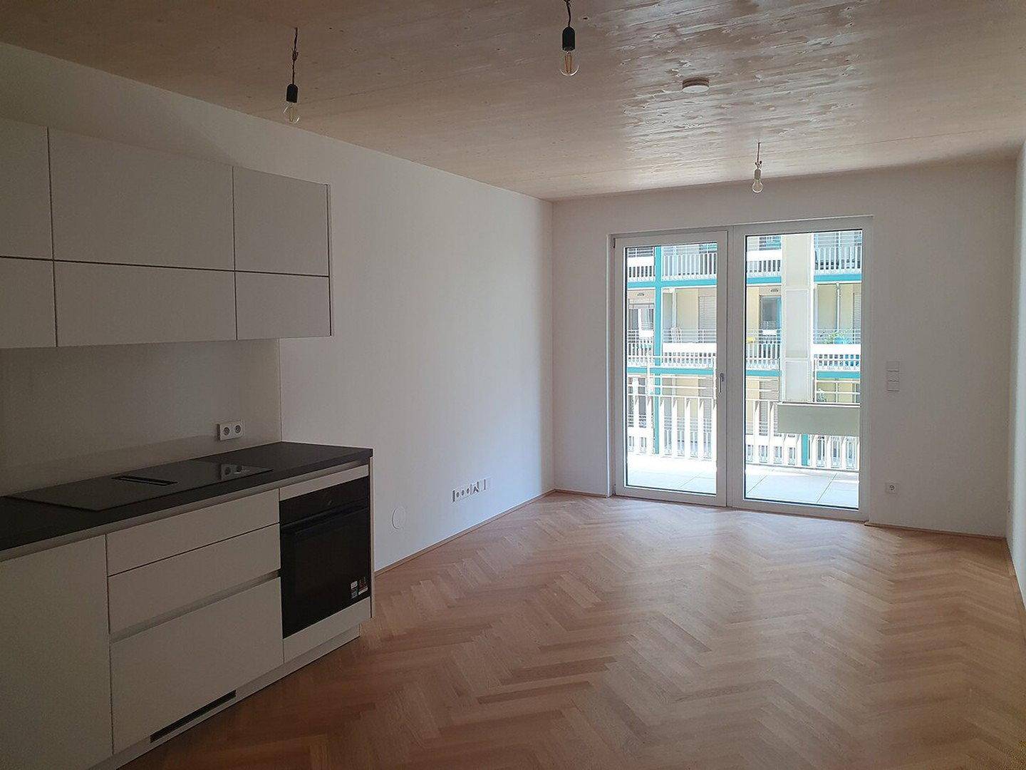 Offener Raum mit moderner Küchenzeile, Fischgrätparkett und Zugang zu einem sonnigen Balkon.