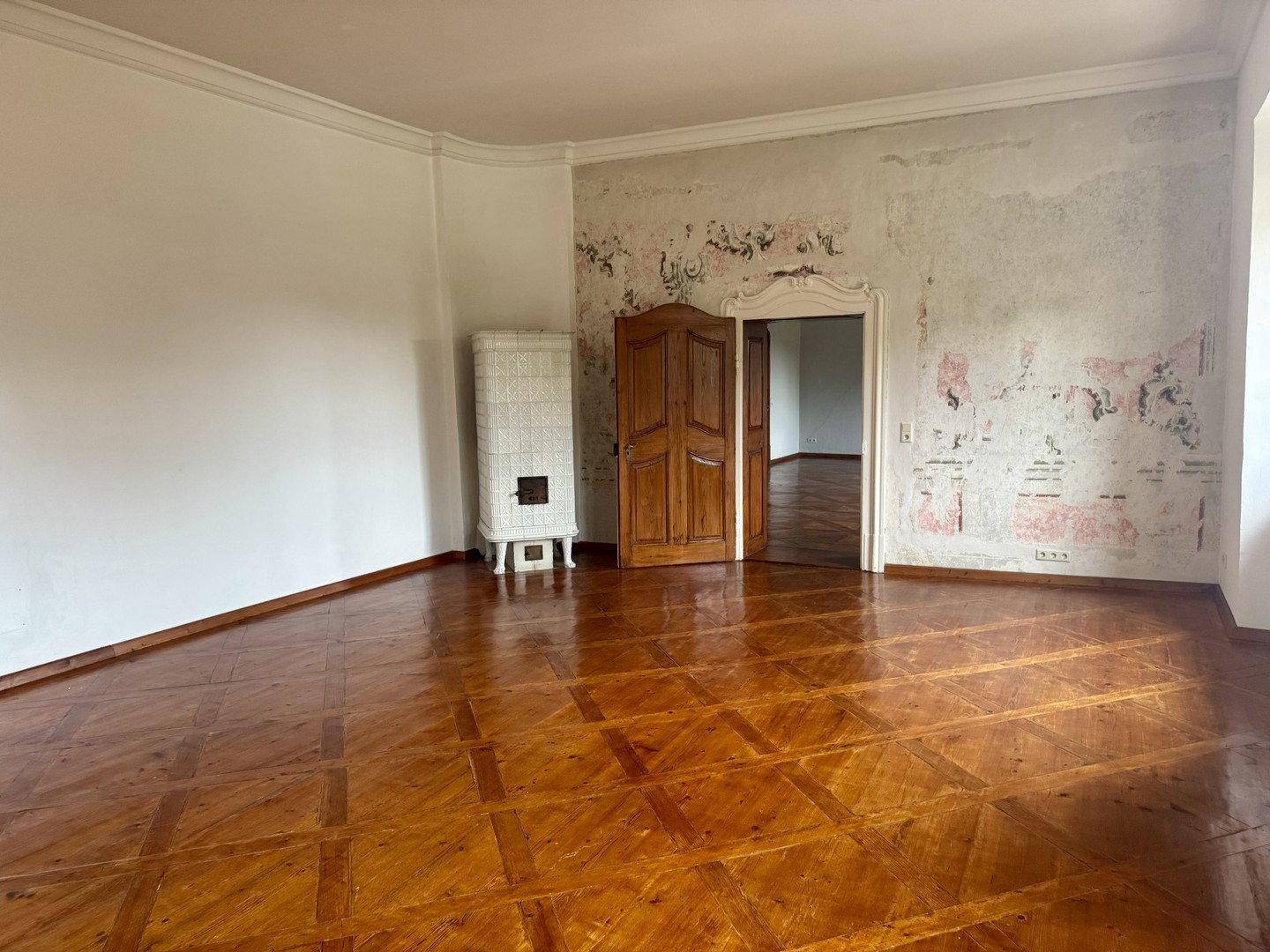 Geräumiger Raum mit Parkettboden und Kachelofen, teilweise renovierungsbedürftige Wand.