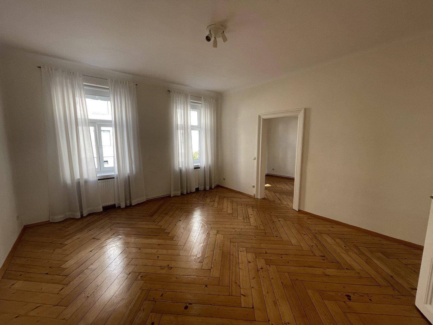Geräumiges Wohnzimmer mit Fischgrätparkettboden, zwei Fenstern und hellen Vorhängen.