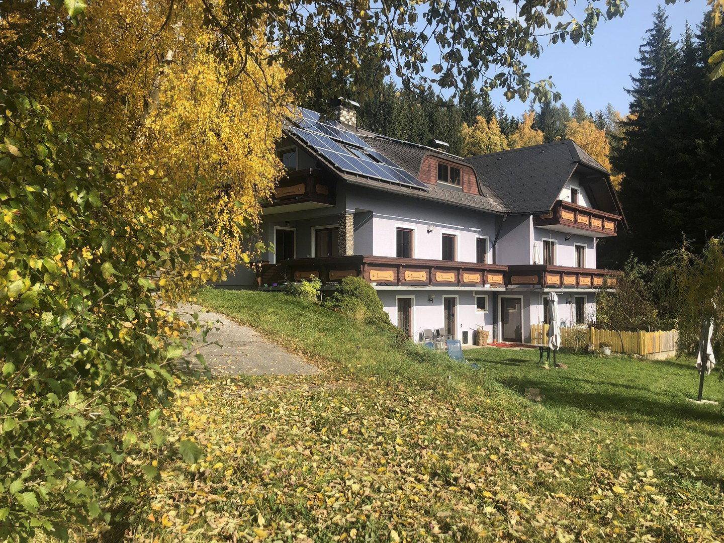 Haus mit Solaranlage und Balkonen, umgeben von herbstlichen Bäumen und Laub.