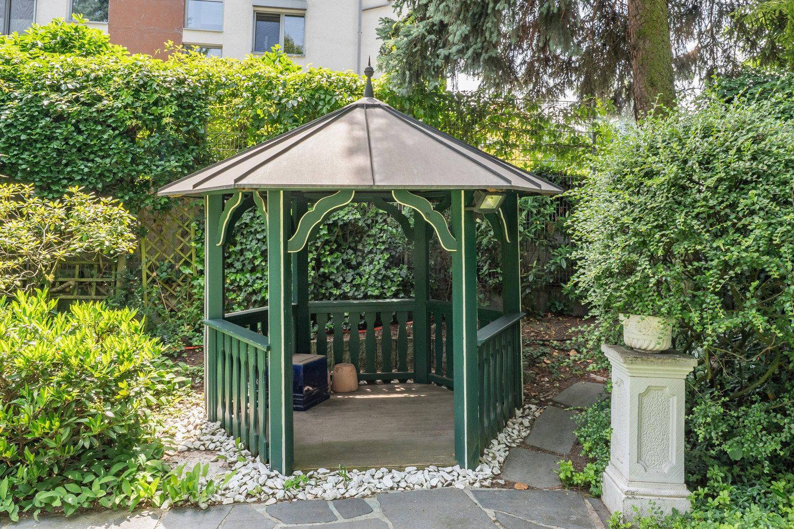 Grüner Gartenpavillon mit Sitzgelegenheiten, umgeben von üppigem Grün und Kies.