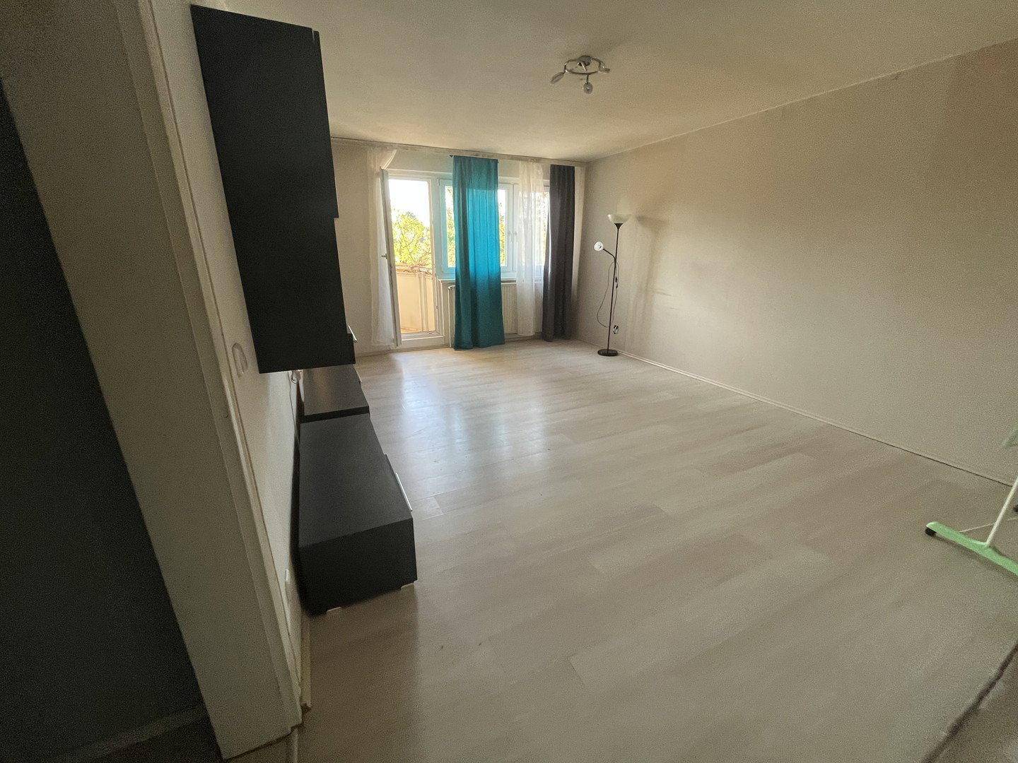 Geräumiges Wohnzimmer mit hellem Laminatboden, dunklem Wandregal und Zugang zum Balkon.