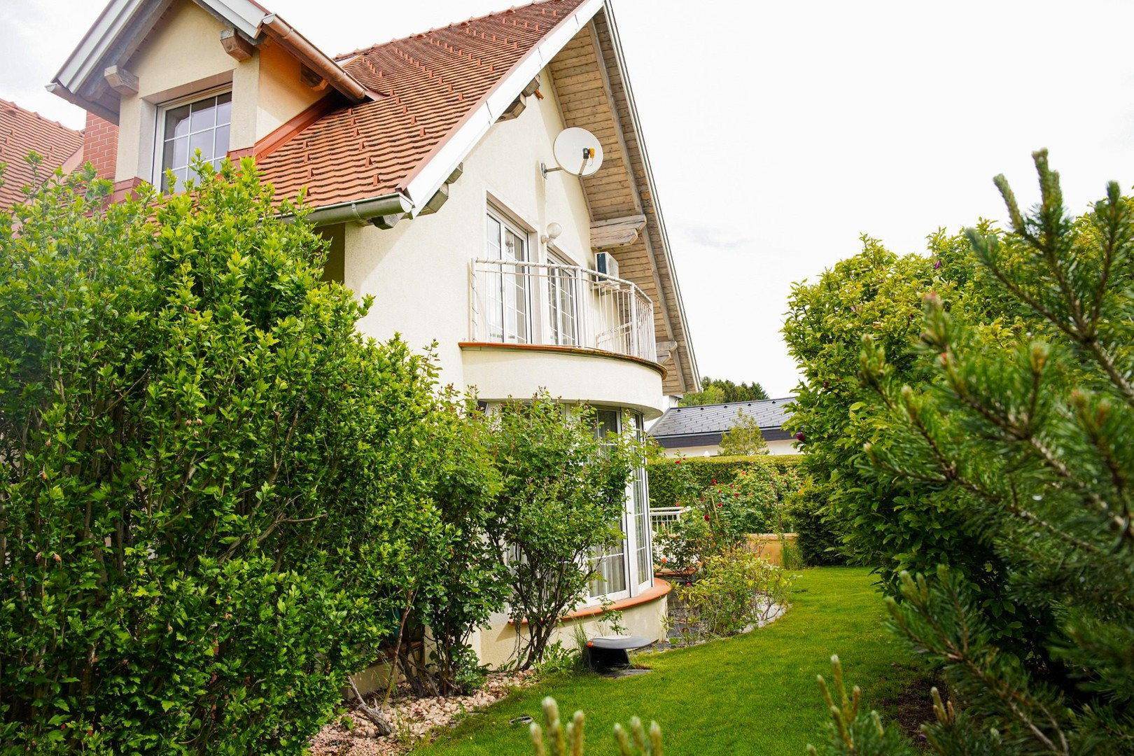 Hausfassade mit Balkon und üppiger Bepflanzung im Vorgarten, unter einem hellen, bewölkten Himmel.