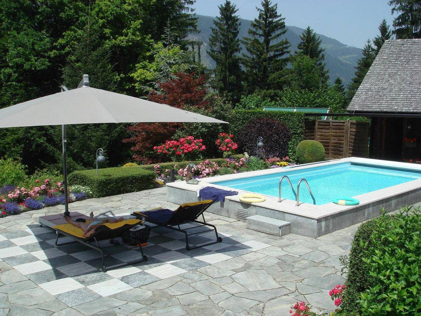 Sonniger Terrassenbereich mit Swimmingpool, Liegestühlen und gepflegtem Garten in den Bergen.