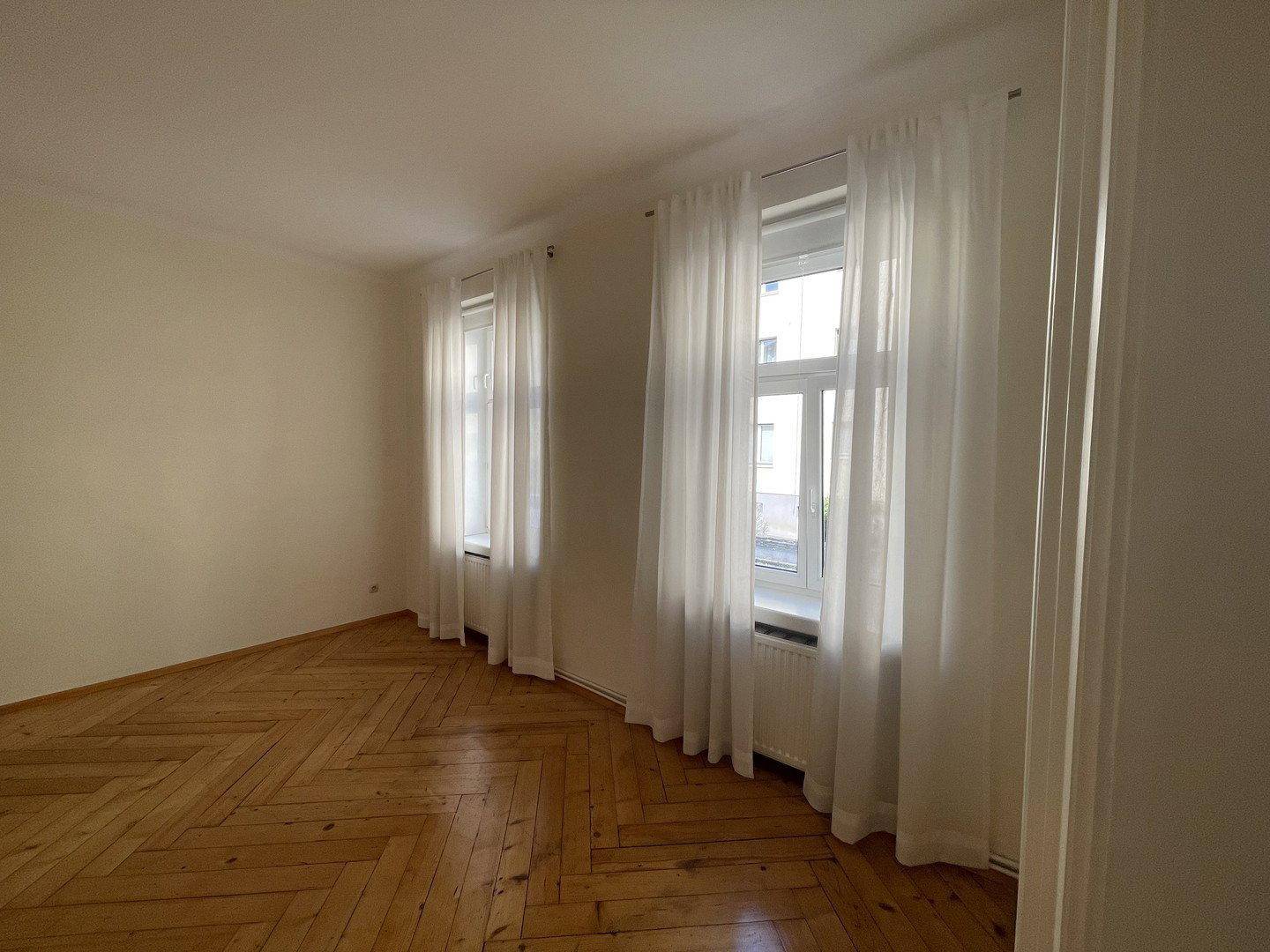 Wohnzimmer mit Fischgrätparkettboden, zwei Fenstern und weißen Vorhängen.
