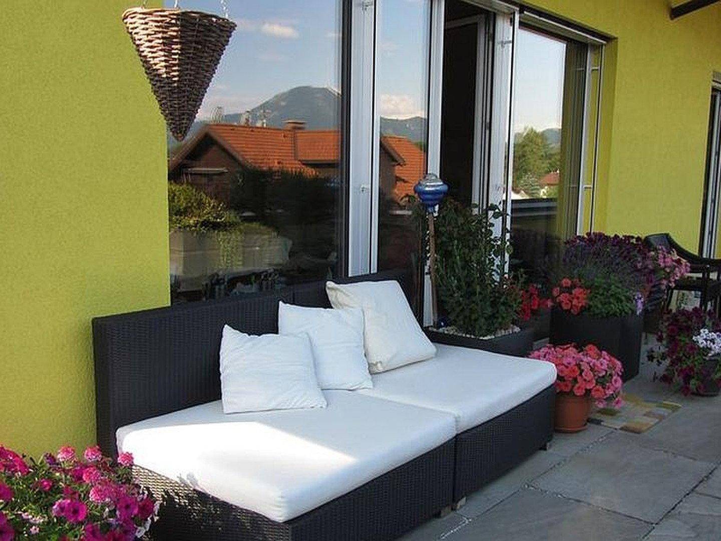 Gemütlicher Balkon mit Rattan-Sofa und weißen Kissen, umgeben von Blumen und mit Bergblick.