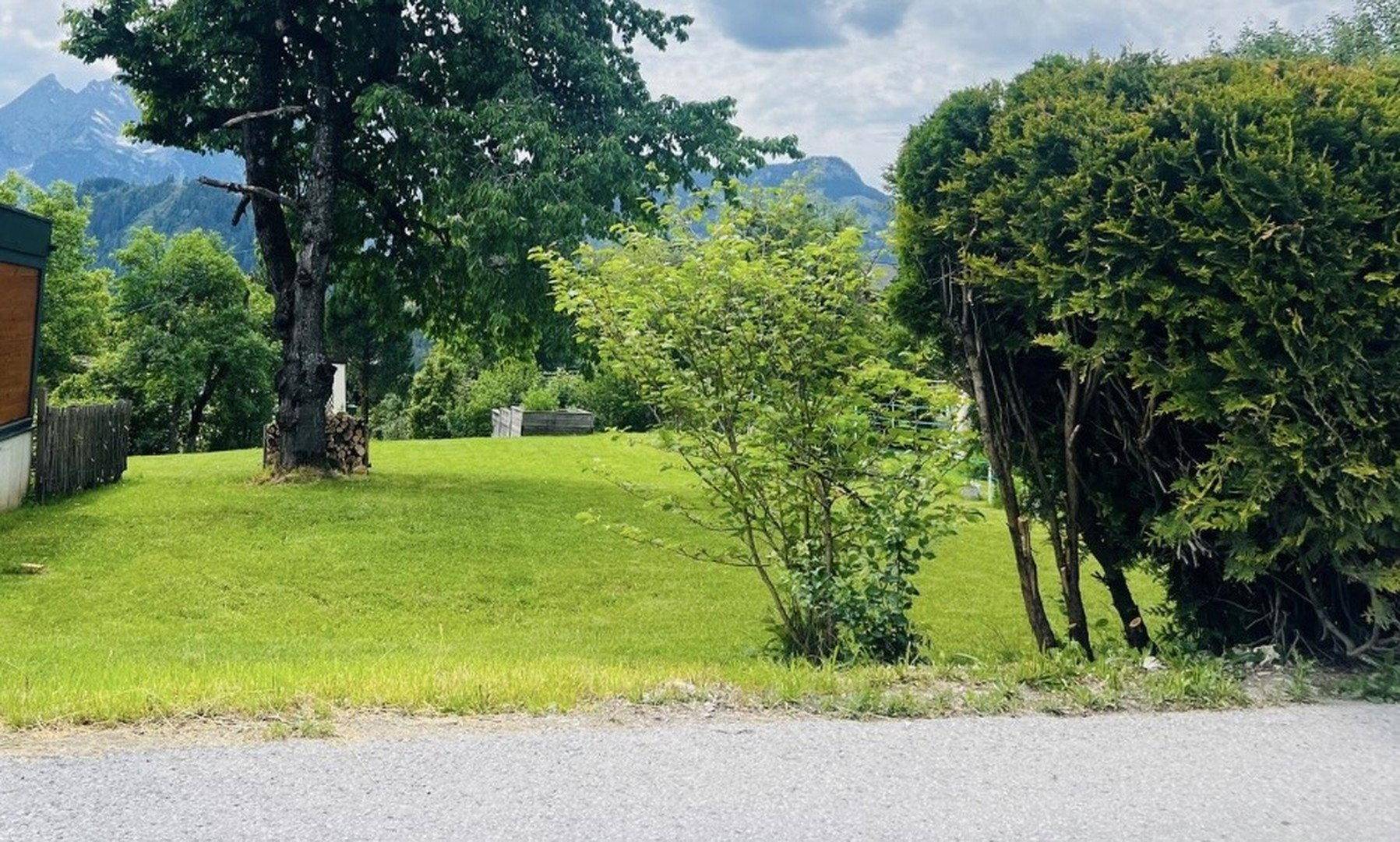 Grüner Garten mit Rasen, Bäumen und Hecken, umgeben von Bergen im Hintergrund.