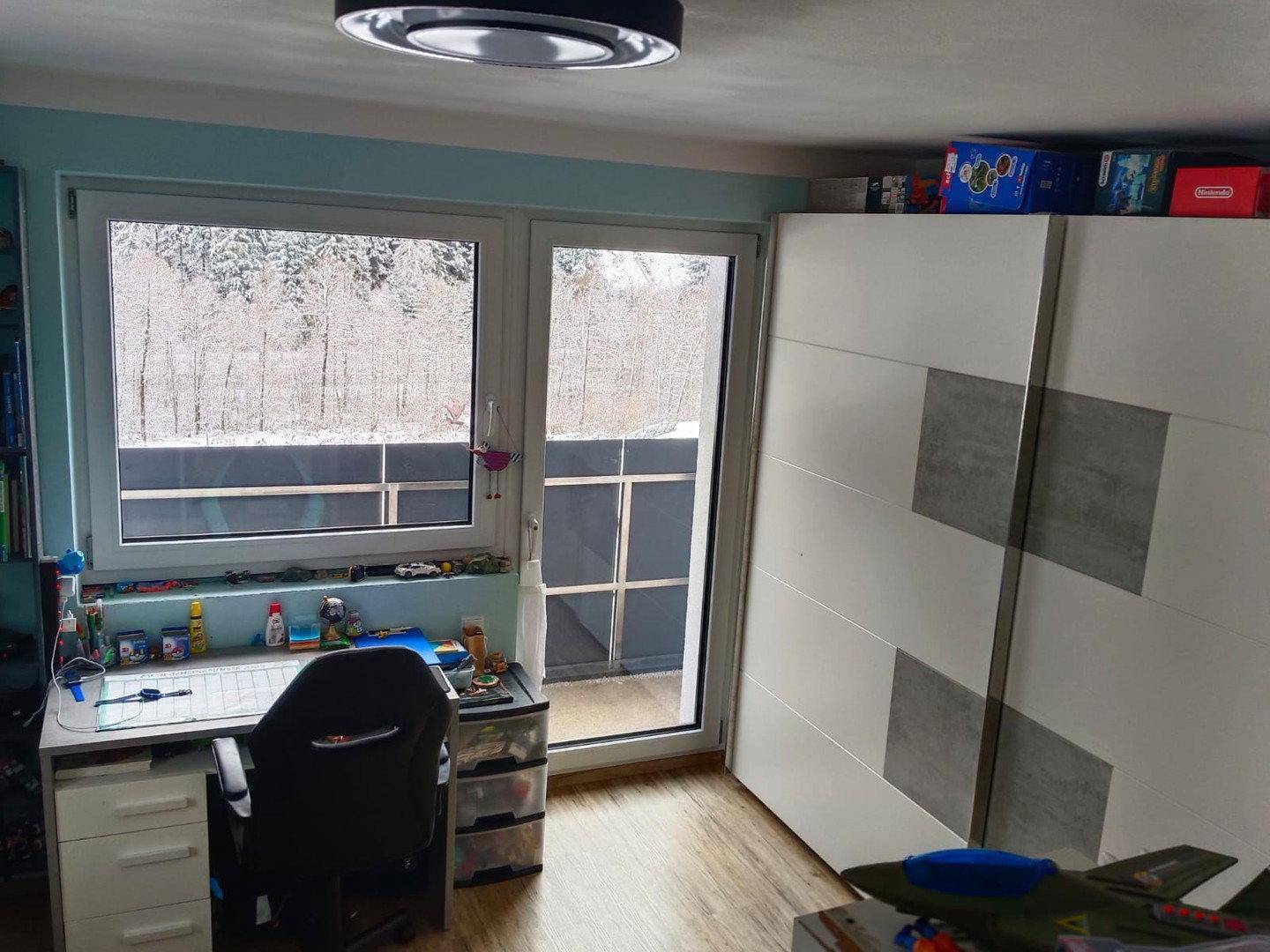 Helles Kinderzimmer mit Schreibtisch, Kleiderschrank und Zugang zu einem Balkon mit Waldblick.