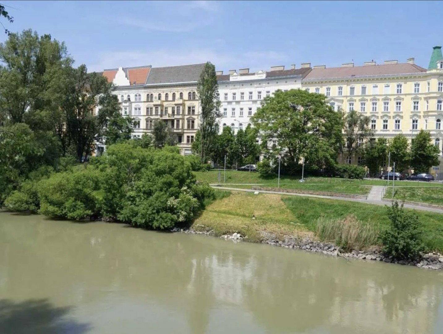 Flussufer mit Grünflächen und einer Reihe klassischer Gebäude im Hintergrund.