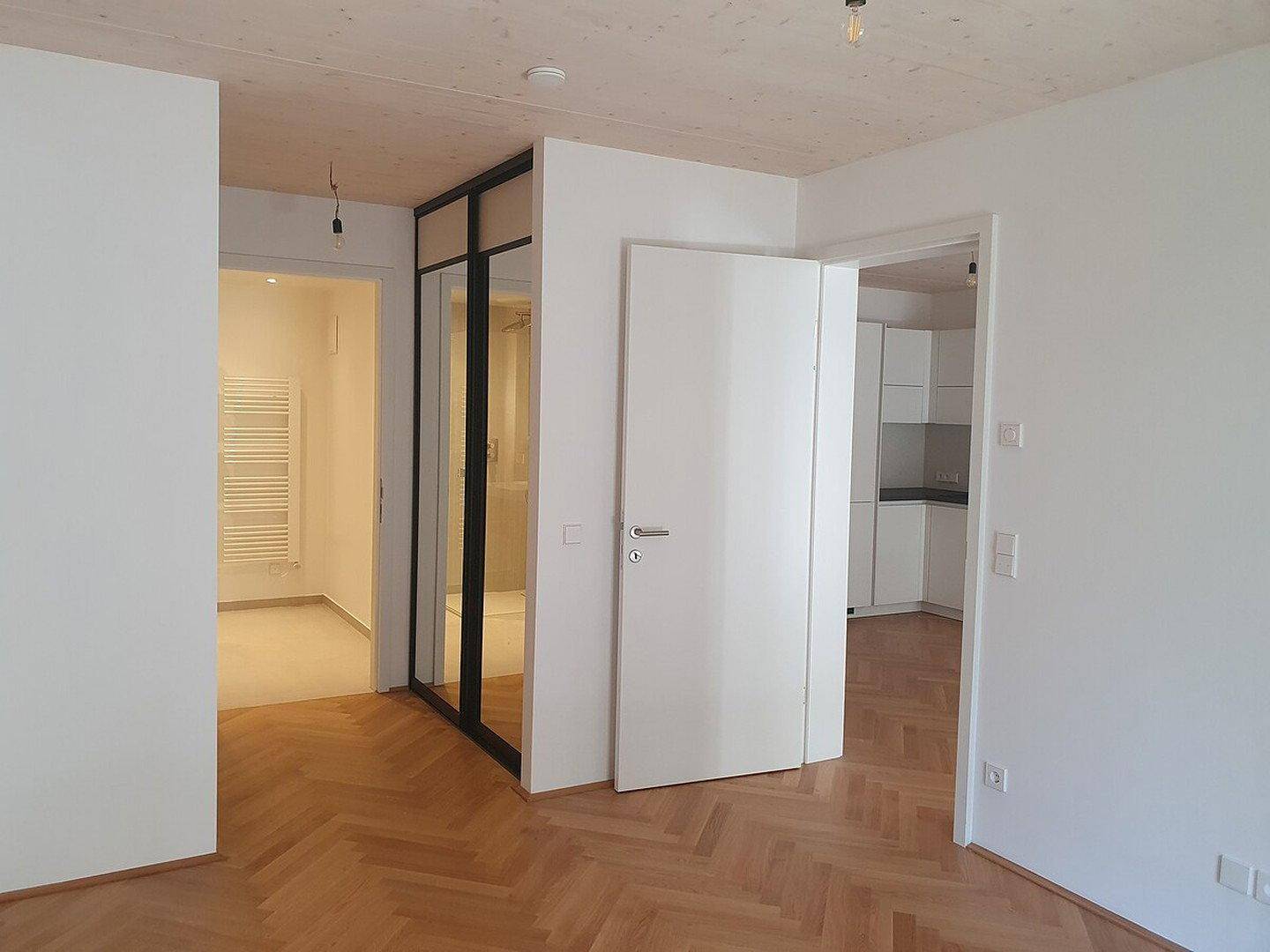 Heller Flurbereich mit Fischgrätparkettboden und Blick in ein modernes Badezimmer mit Glasdusche.