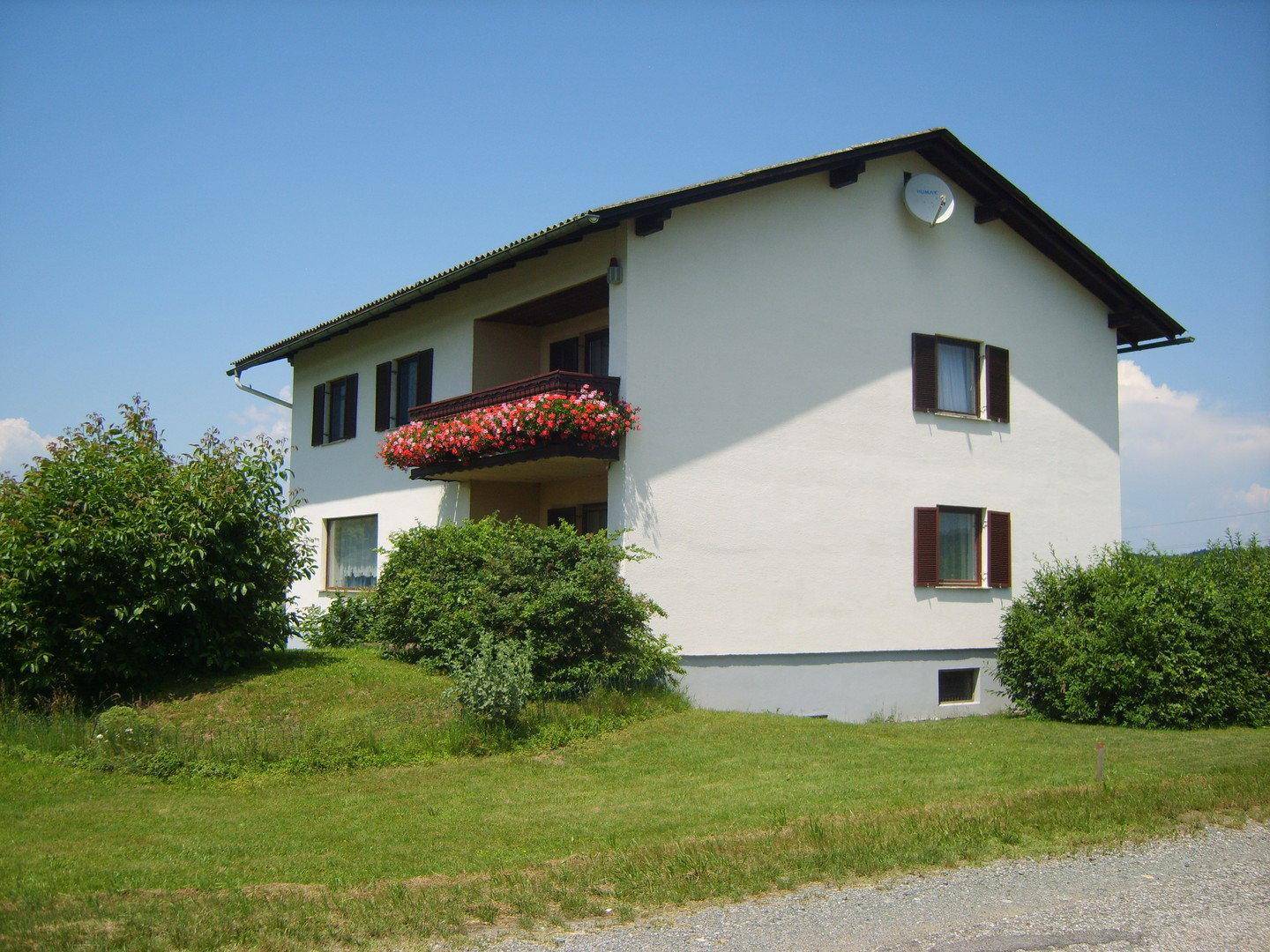 Weißes Wohnhaus mit braunem Dach, Fensterläden und einem blumengeschmückten Balkon in ländlicher Umgebung.