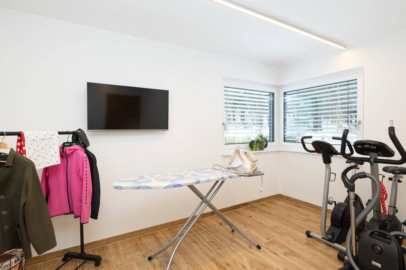 Mehrzweckraum mit Fitnessgeräten, Bügelbrett und Fernseher, auch als Hauswirtschaftsraum nutzbar.