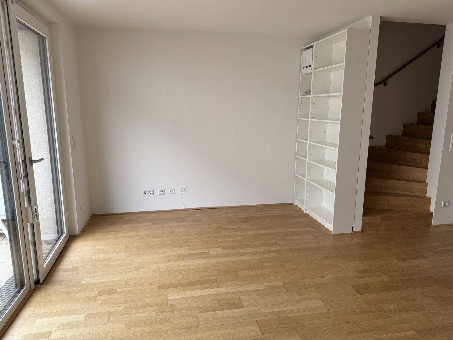 Heller Raum mit Holzfußboden, weißem Regal und Zugang zu einem Balkon, ideal als Wohnbereich.