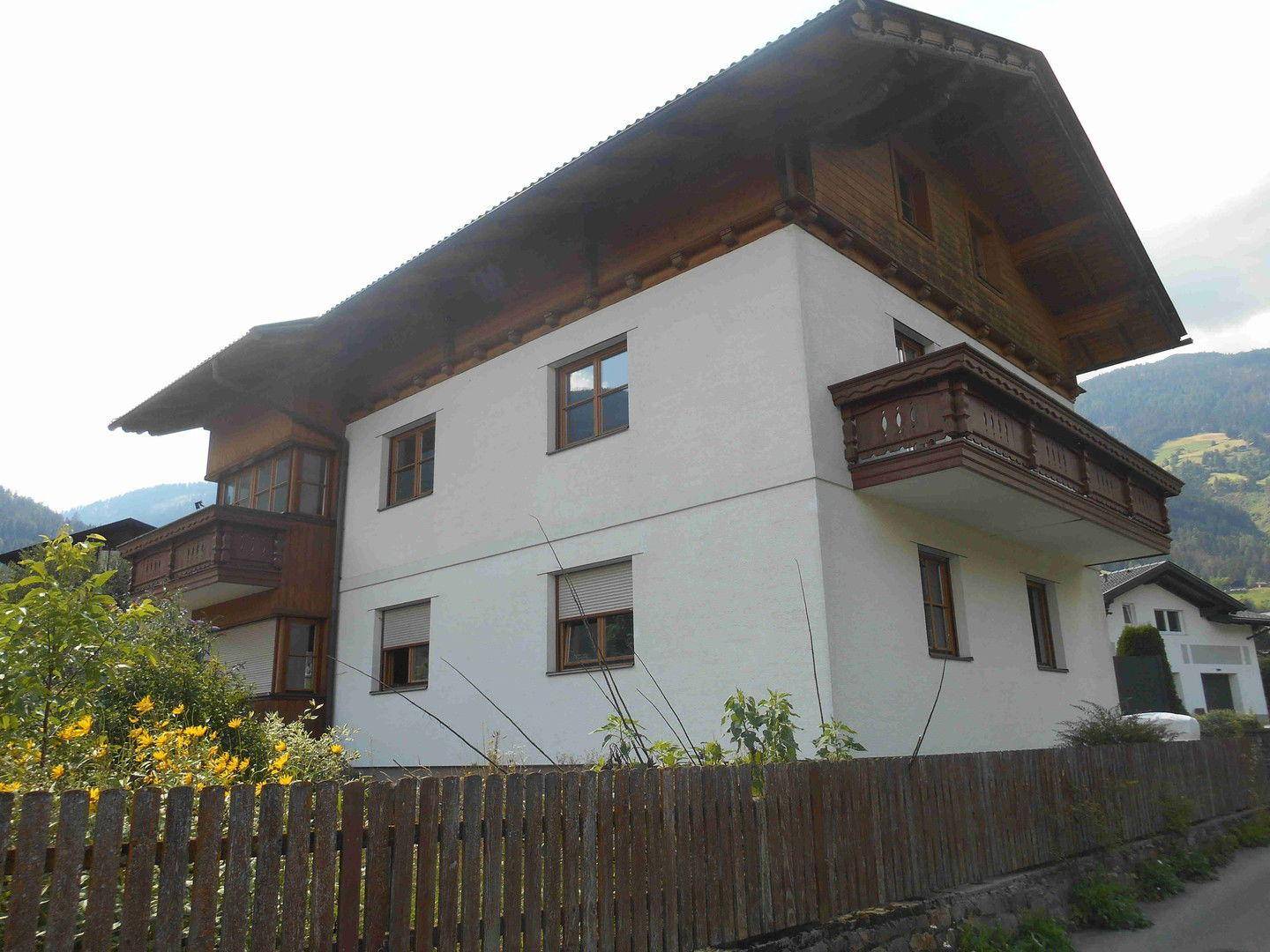 Seitenansicht eines traditionellen Hauses mit Holzbalkonen und weißer Fassade vor bergiger Kulisse.