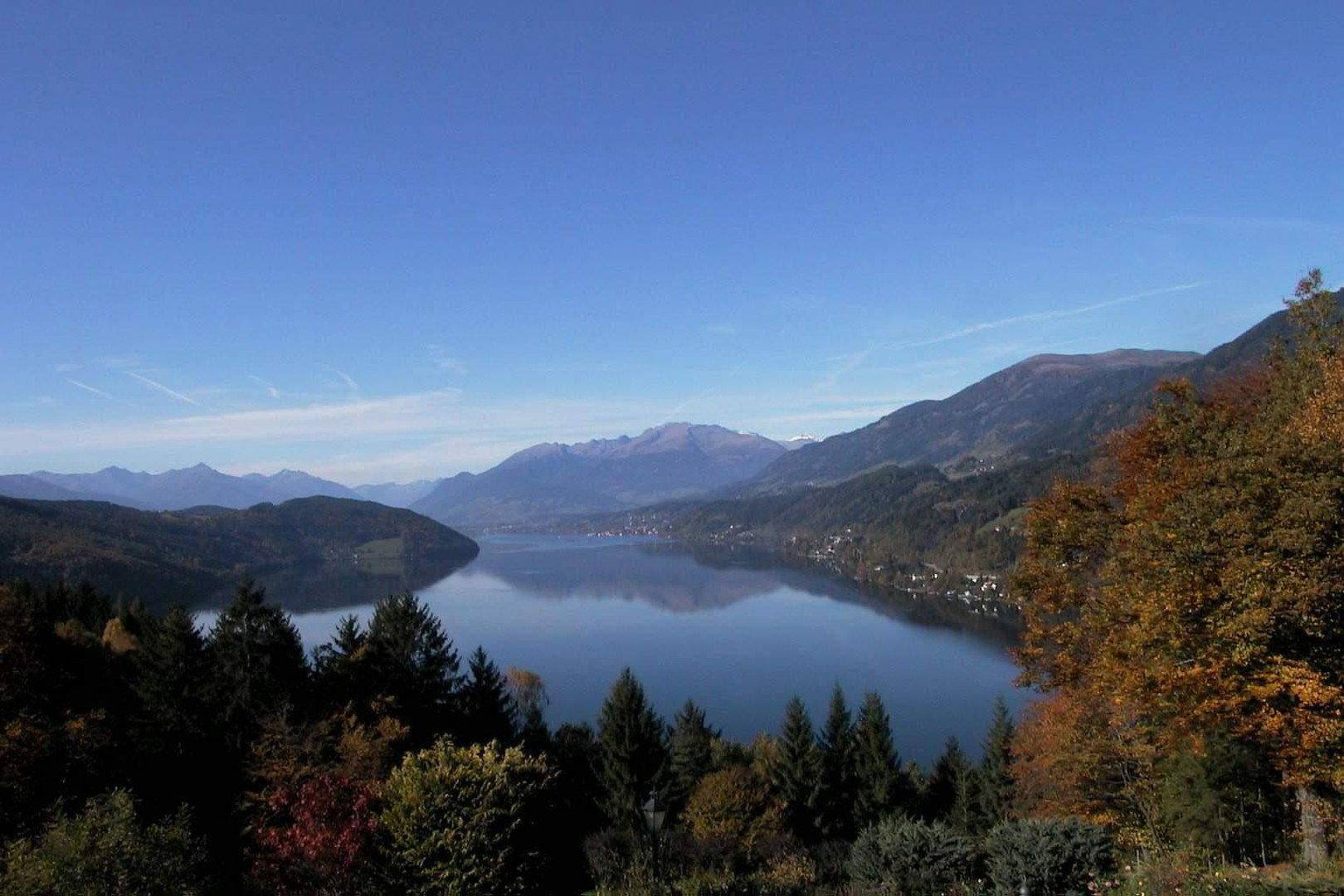 Weitläufiger Blick auf einen ruhigen See, umgeben von bewaldeten Hügeln und Bergen unter blauem Himmel.
