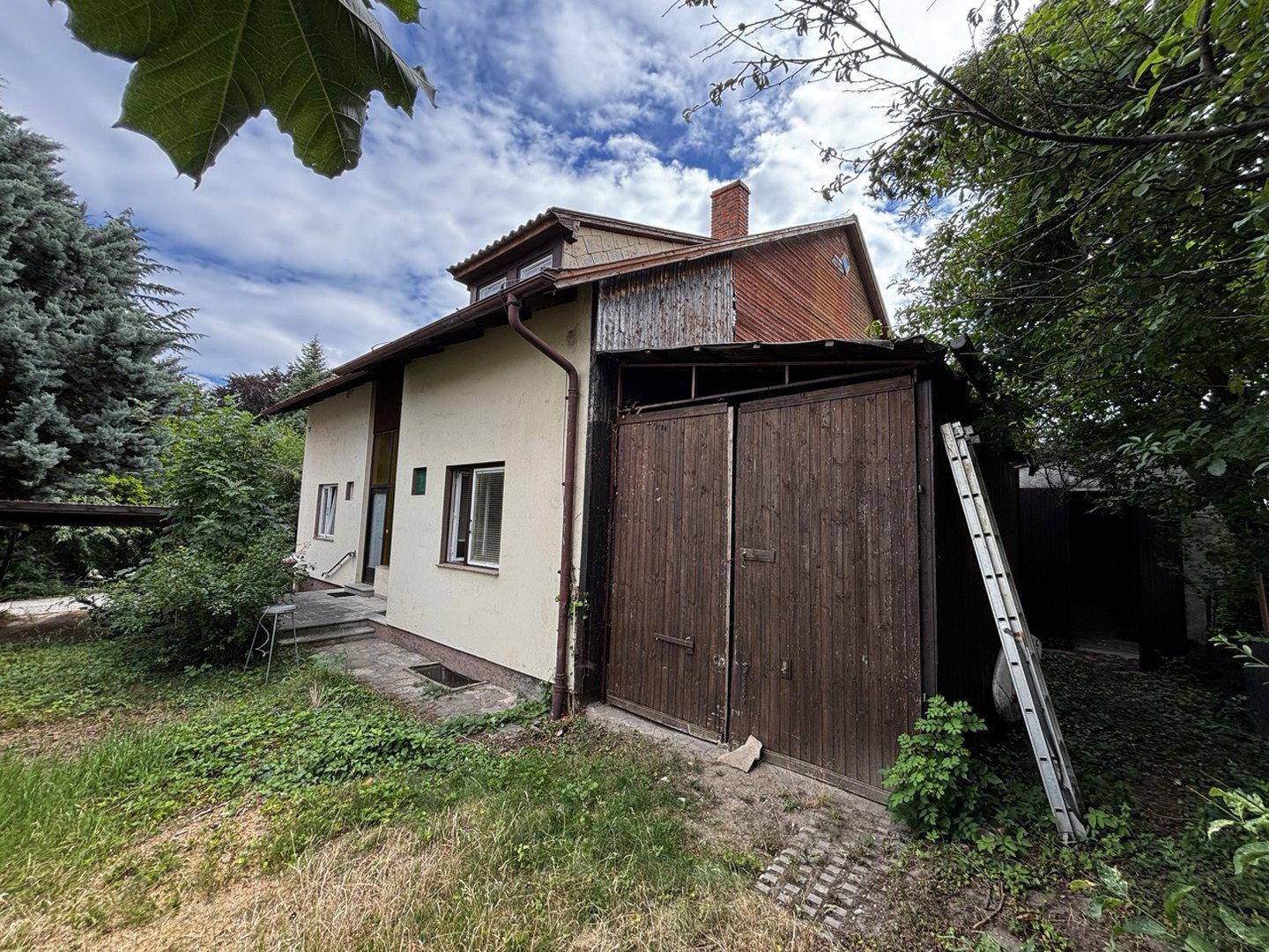 Hausfassade mit angrenzender Holzgarage und umgebendem Grün.