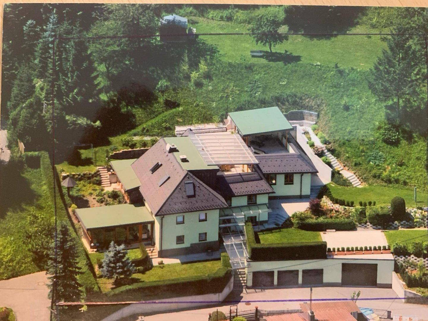 Luftaufnahme des Anwesens mit mehreren Gebäuden, Gartenflächen und Carport in grüner Umgebung.