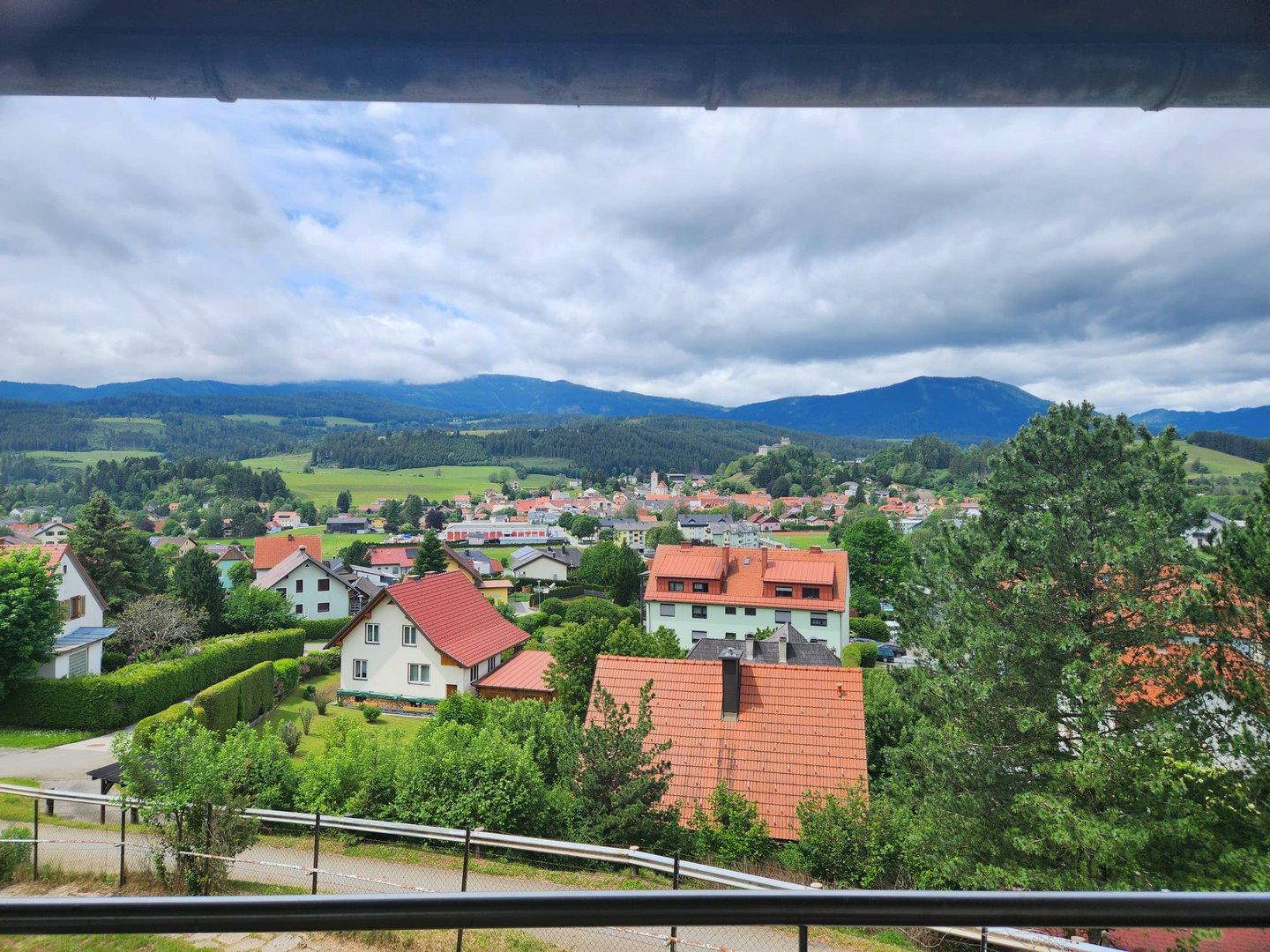 Weitblick vom Balkon über ein Dorf, grüne Hügel und eine malerische Berglandschaft in der Ferne.
