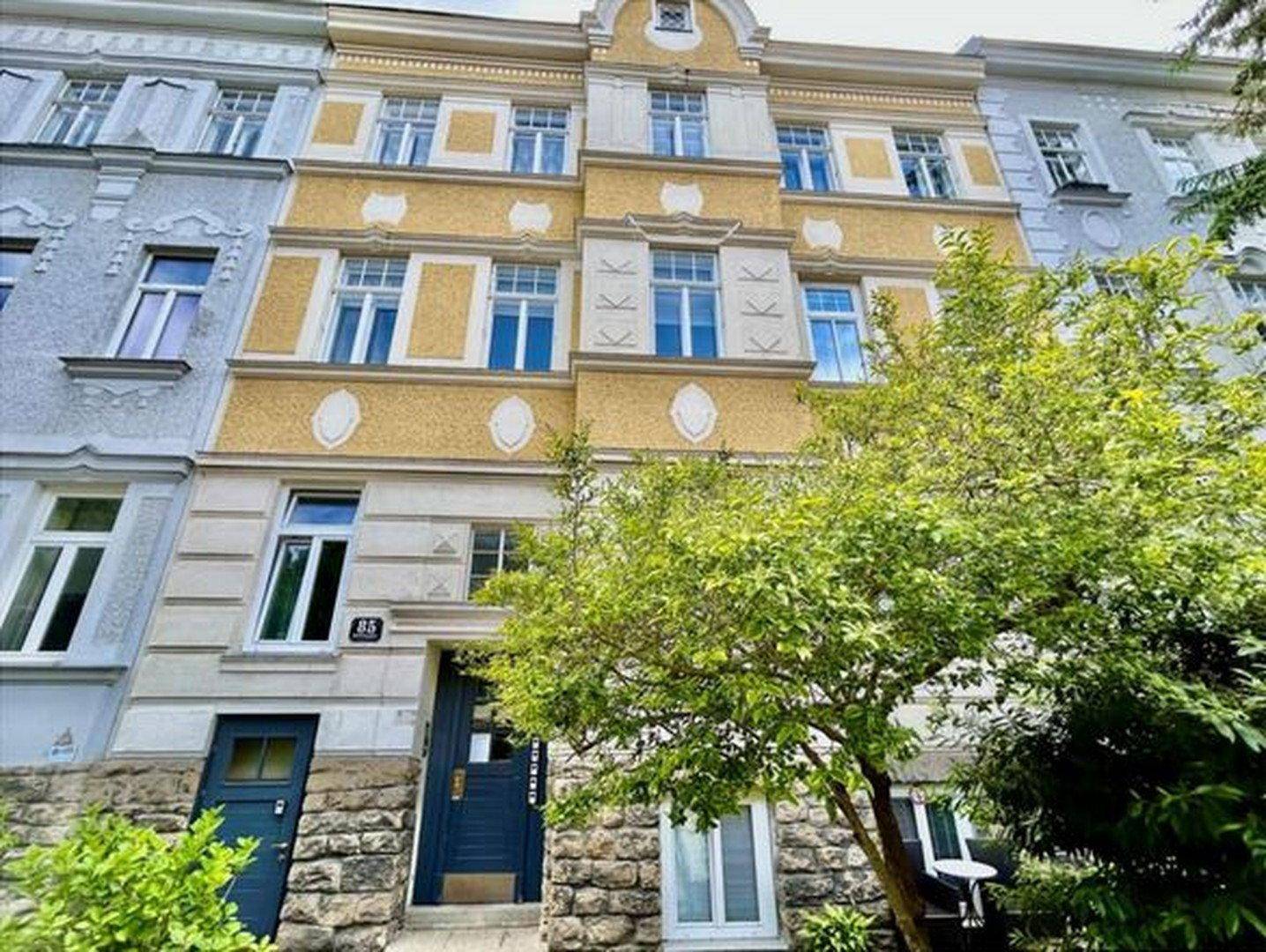 Charmantes Altbaugebäude mit gelber Fassade, verzierten Fenstern und einem Baum im Vordergrund.