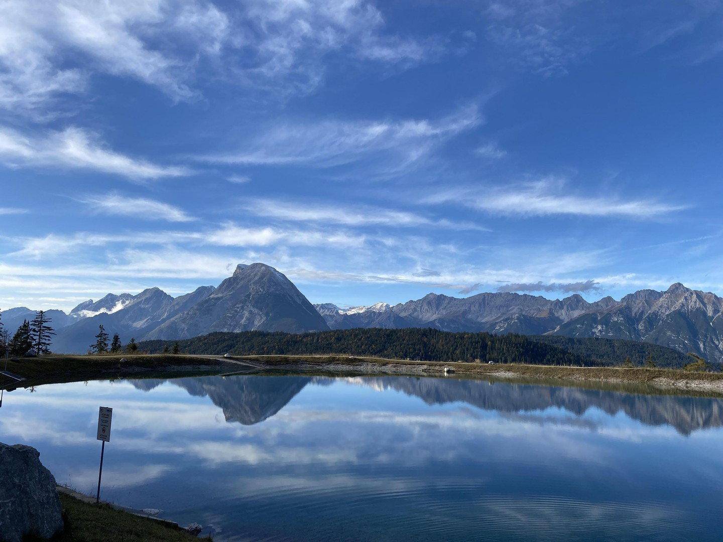 Klarer Bergsee mit spiegelnder Oberfläche und majestätischen Gipfeln unter blauem Himmel.